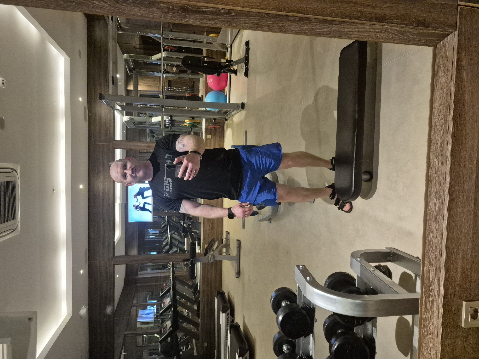 A reflection in a mirror shows a man in a gym. The man is taking a selfie with his phone. He is wearing a black t-shirt and blue shorts. He also has a smartwatch on his left wrist. The gym contains various workout machines, dumbbells, and a weight bench. In the background, there are several television screens and wooden wall panels. The gym's location is in Alanya, Turkey.