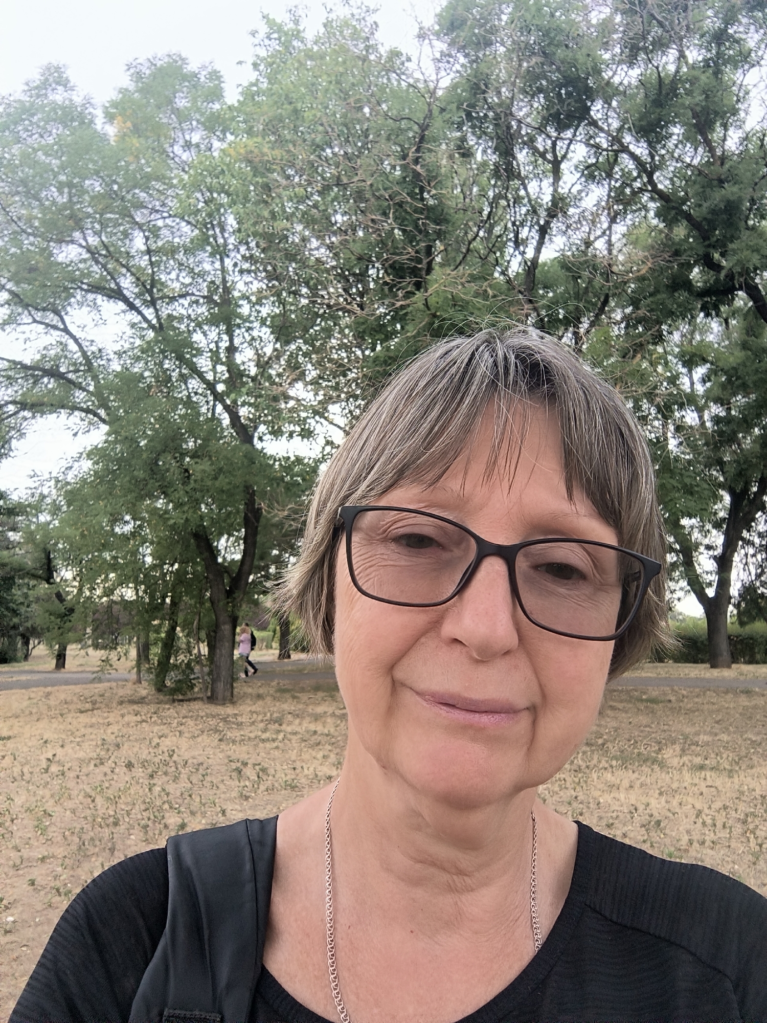 A selfie is taken by a woman with short, graying brown hair and glasses. She is wearing a black top and a silver necklace. She is standing outdoors in a park-like setting with trees and dry grass. In the background, two people can be seen walking.