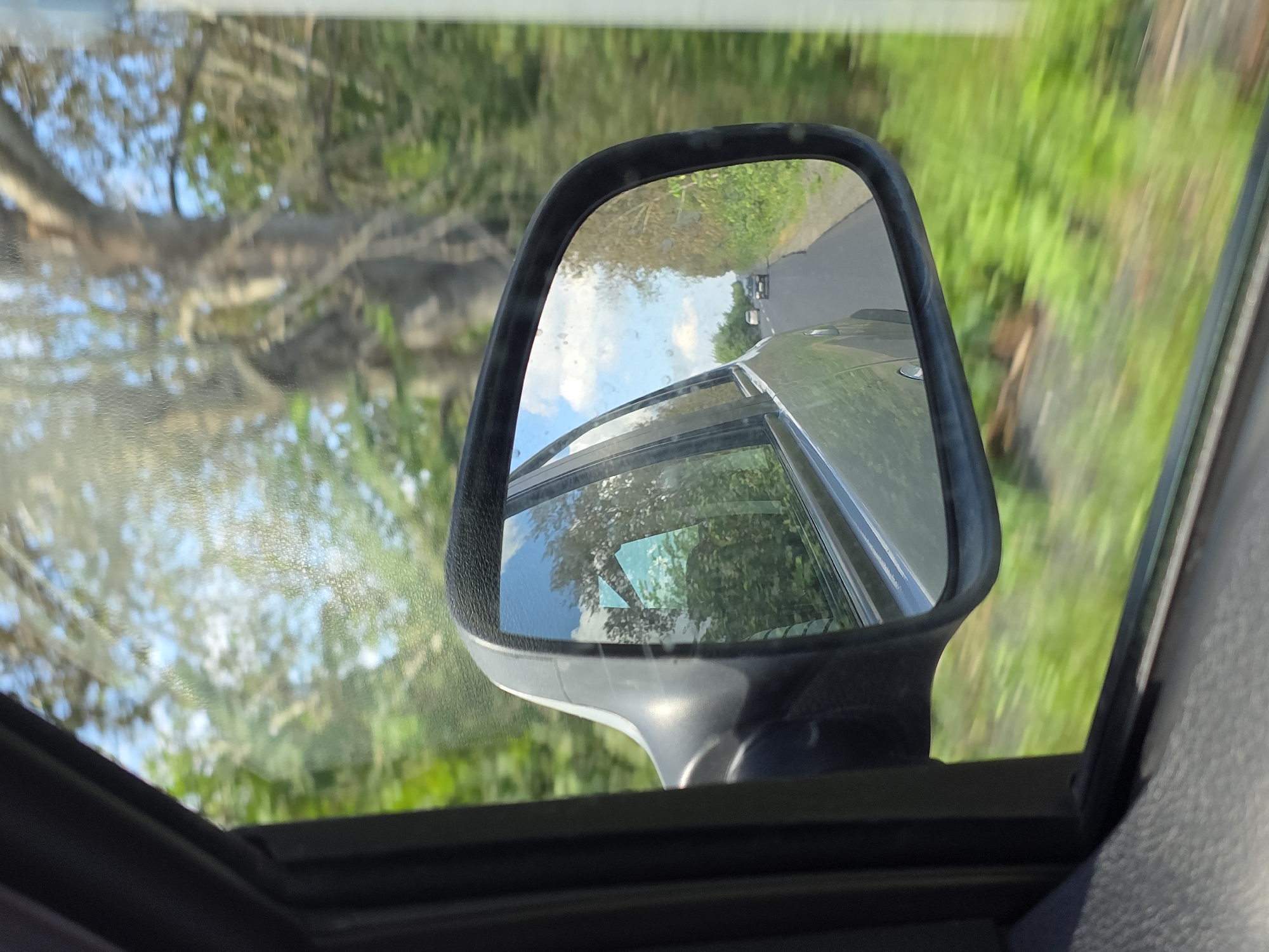 The image shows a rear-view mirror of a car, reflecting the road and surrounding environment. The mirror shows a grey car in front, driving on a road bordered by green trees and vegetation. The sky is blue with white clouds. The scene appears to be a rural or suburban area. There are no people visible. The location is identified as Yaratkulova, Russia.