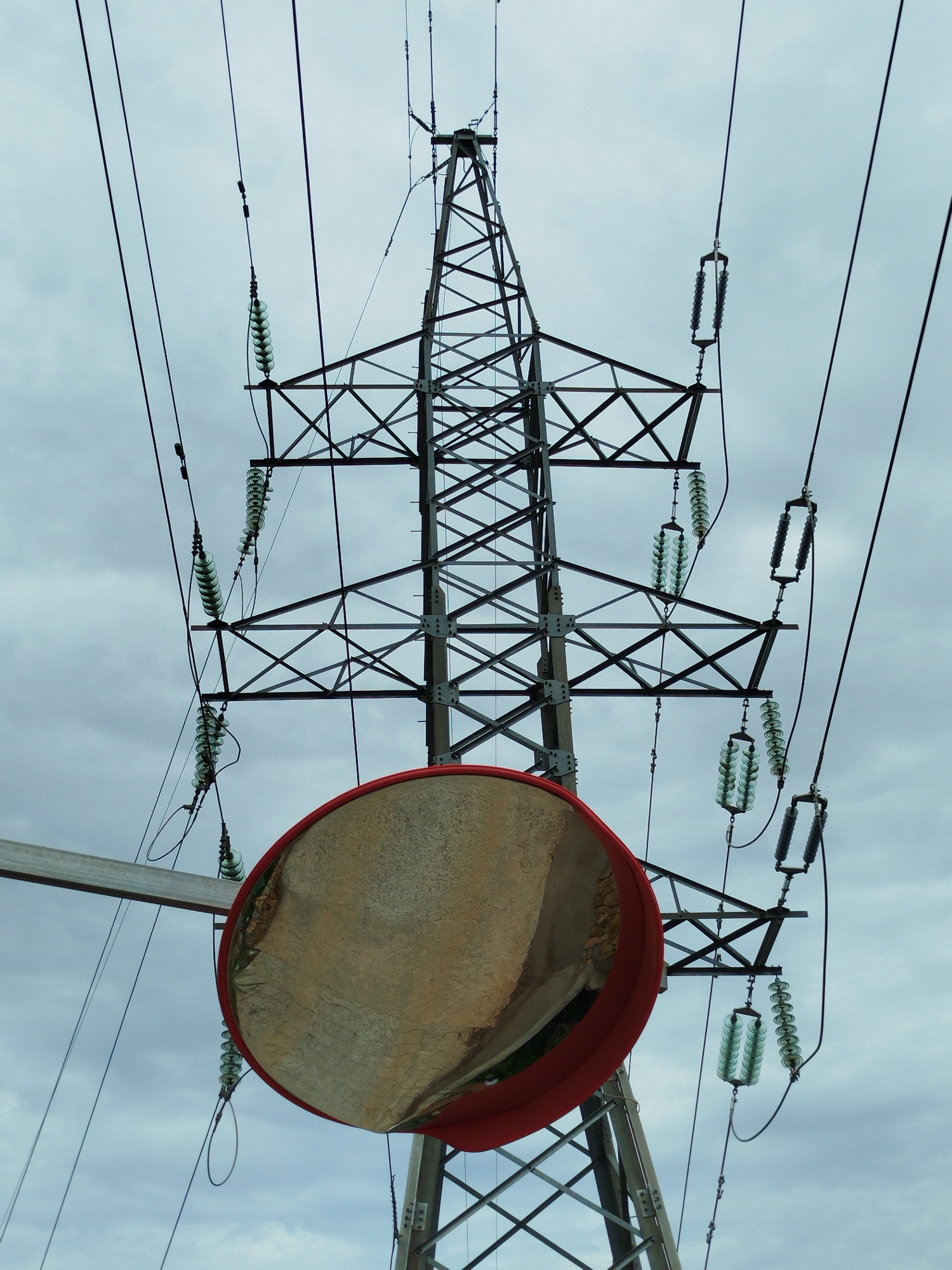 A low-angle, full-shot image reveals an electrical transmission tower situated in Da Lat, Vietnam. The tower is constructed of metal lattice and supports high-voltage power lines via glass insulators. A large, circular, red-rimmed convex mirror is affixed to the tower's structure at a low level. The sky in the background is overcast. No people are visible.