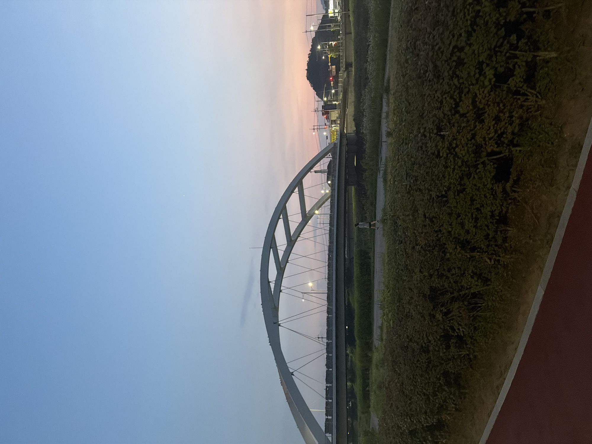 This is an outdoor, eye-level, medium shot photograph of the Somang Bridge in Dongducheon-si, South Korea, taken at dusk. The sky displays a gradient of light blue at the top transitioning to soft pink and orange hues closer to the horizon. The Somang Bridge, a large white arch bridge, dominates the mid-ground. Its structure is composed of a curving arch supported by numerous diagonal cables. The bridge spans over a dark, calm body of water, likely a river, which is partially obscured by lush green bushes in the foreground. A pedestrian can be seen walking on a path to the right of the bridge. The background features a hill topped with buildings and a network of streetlights, indicating an urban or semi-urban setting. The foreground consists of a dark red path, possibly a running or cycling track, bordered by a narrow strip of dirt.