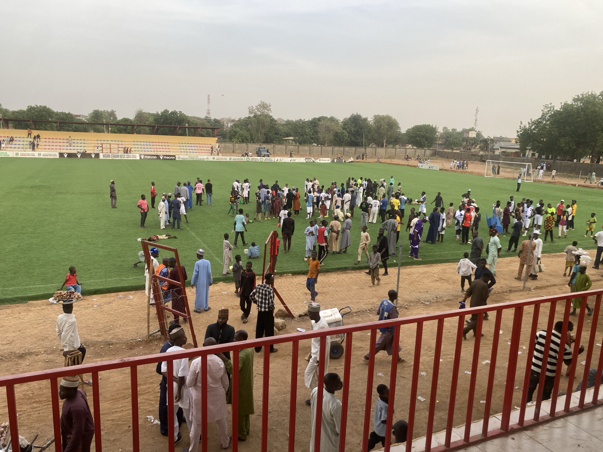 The image depicts a large gathering of people on and around a green soccer field. The field is surrounded by stands, with a yellow section visible on one side. A red railing is visible in the foreground, suggesting the image was taken from a slightly elevated position. The people appear to be diverse in age and attire, with many wearing traditional clothing. There are people standing, walking, and some are interacting in small groups. A few individuals are near a gate in the foreground. The setting implies the location is in or near a stadium, possibly in Tudun Wada, Nigeria. The sky is overcast, suggesting the image was taken on a cloudy day.
