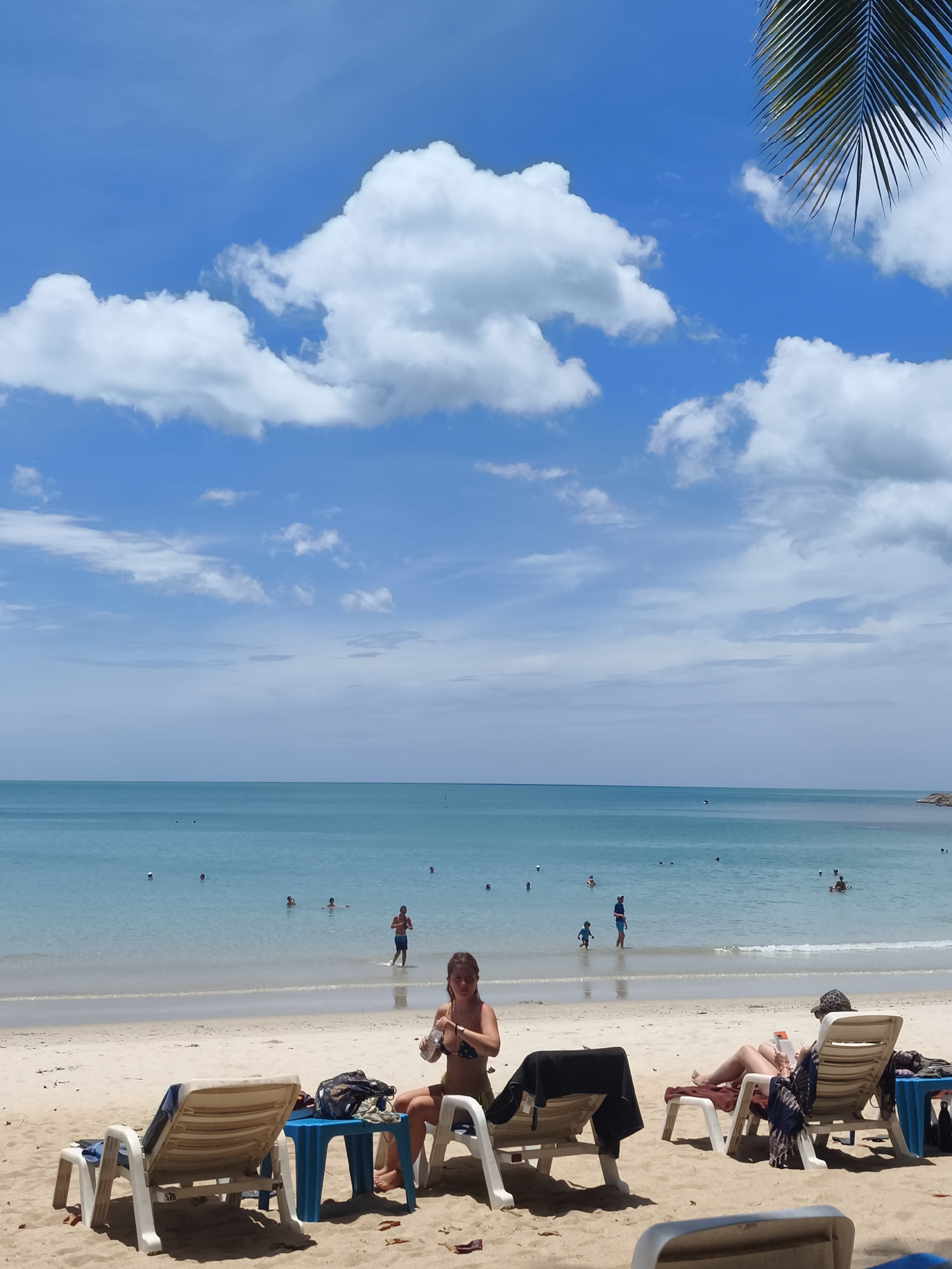 In the city of Ko Samui District, Thailand, a tranquil beach scene unfolds under a bright blue sky dotted with fluffy white clouds. The turquoise water of the ocean gently laps at the sandy shore. Several people are visible in the shallow water, some wading and others swimming.
In the foreground, two beach chairs sit on the sand. On the right, a woman wearing a bikini is seated in a lounge chair, holding a bottle of water. Next to her, a blue side table holds a bag. To the left, another lounge chair is empty, with a black towel draped over it. Further into the water, a man in blue shorts stands, and a small child walks towards him. In the distance, more people can be seen enjoying the water. The overall atmosphere is one of relaxation and enjoyment of a sunny day at the beach.