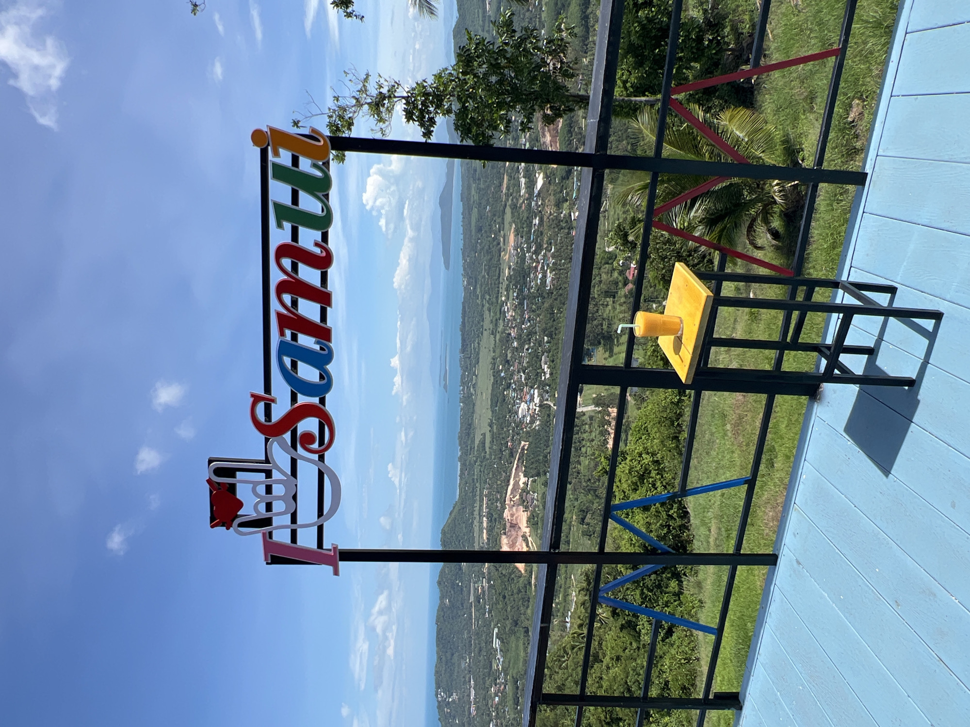 The image depicts an elevated outdoor viewpoint overlooking a vast coastal landscape under a clear blue sky with scattered white cumulus clouds. In the upper mid-ground, a prominent sign reads "I ❤️ Samui". The "I" is pink, followed by a white stylized hand gesture forming a 'love' symbol with a red heart at its apex. The word "Samui" is rendered in multi-colored, curved block letters: a red "S", blue "a", green "m", red-orange "u", and orange "i" with an orange dot. The sign is supported by black vertical posts.
In the lower right foreground, an orange beverage with a white straw rests in a clear glass on a yellow square stool with black legs. A black metal railing with horizontal bars spans the width of the view, featuring integrated decorative triangular supports: blue on the left and red on the right, resembling stylized 'M' or 'A' shapes. The platform floor in the immediate foreground consists of light blue painted wooden planks.
Beyond the railing, an expansive panorama unfolds. The mid-distance reveals a lush green landscape of rolling hills and valleys, densely covered in trees and vegetation. Scattered among the greenery are numerous residential and commercial buildings, along with a prominent, large, light brown quarry excavation site. In the far distance, the serene turquoise sea meets the horizon, dotted with several distant islands and smaller landmasses. No people are present in the scene. This location is situated in Baan Thurian, Thailand.