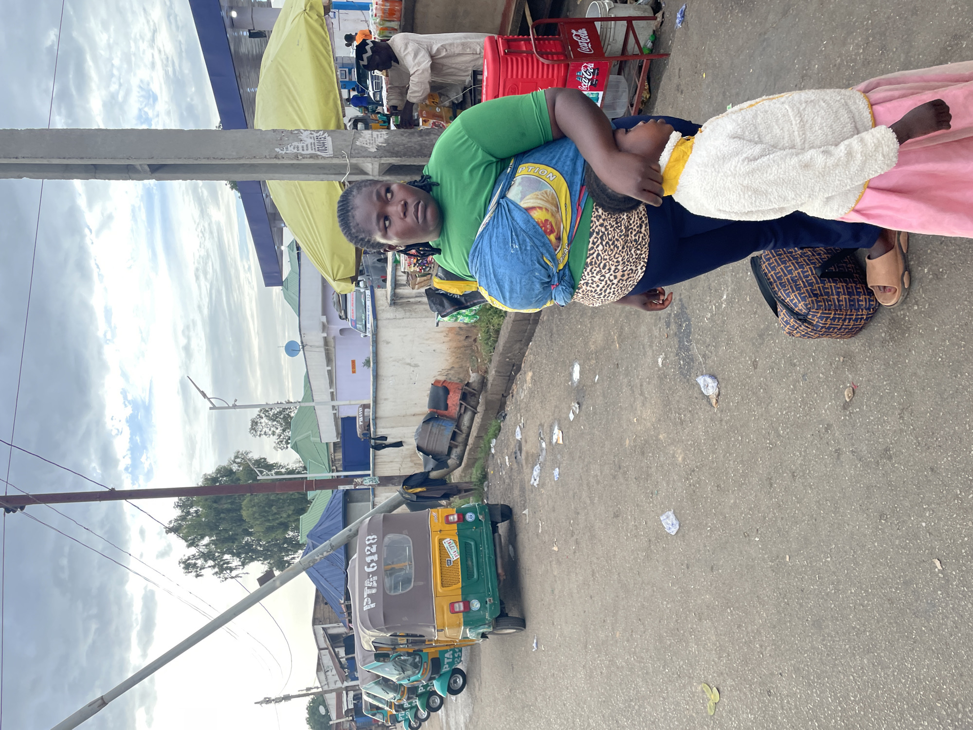 The image shows a woman carrying a child on her back in an outdoor location. The woman wears a green shirt, dark pants, and sandals, and carries a blue patterned bag. The child is wearing a yellow top, a white fuzzy jacket, and a pink skirt. The child is being carried on the woman's back. In the background, there are auto rickshaws lined up, a yellow tent-like structure, a Coca-Cola cooler, and people in the distance. Debris is visible on the ground. The picture was taken during the daytime under cloudy skies in what appears to be a street scene in Jos, Nigeria. The auto rickshaws parked in the background are a common form of transportation in the city.