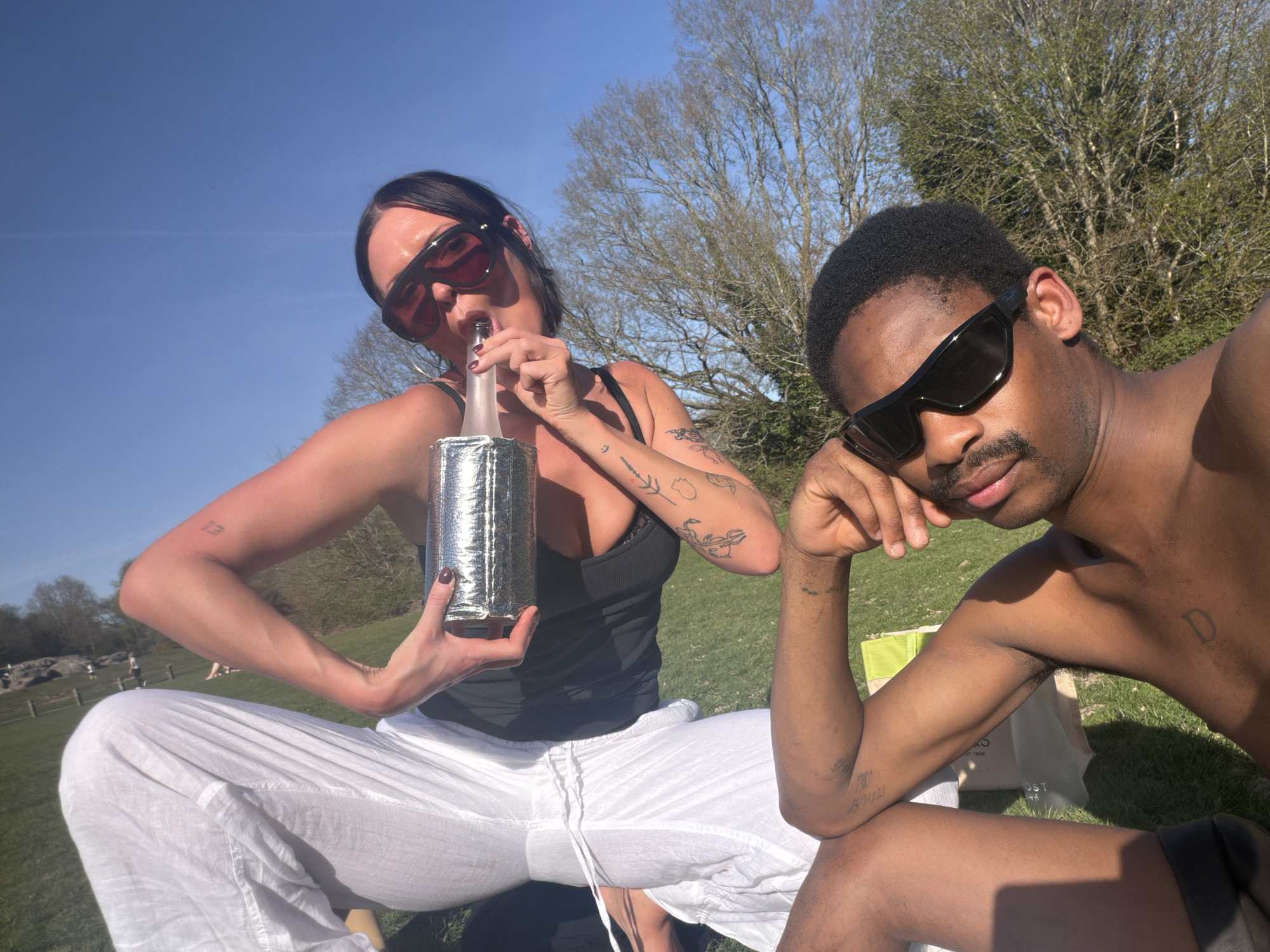Two people are sitting on a grassy field under a clear blue sky with sparse trees. The person on the left is a woman with dark hair, wearing sunglasses, a black tank top, and white pants. She holds a bottle covered in reflective material to her lips as if drinking from it. She has multiple visible tattoos on her arms. To her right is a Black man wearing sunglasses and appearing to be shirtless. His head is resting on his hand. He also has tattoos on his arms. The background shows more grassy fields, trees, and a few people in the distance. The location is Tunbridge Wells, United Kingdom.