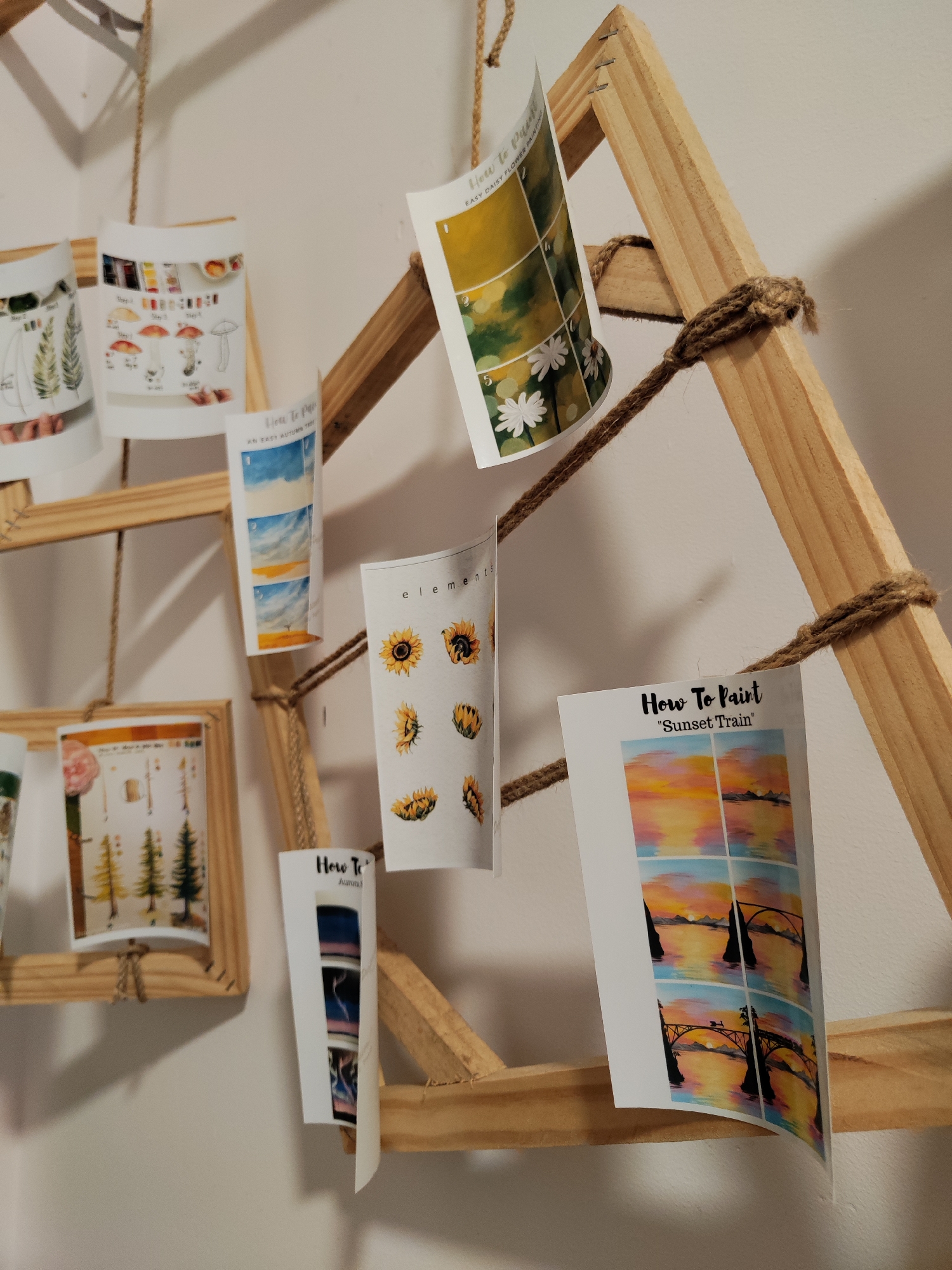 A close-up shot features a collection of printed art instructions hanging from a wooden A-frame structure, likely indoors against a plain white wall. The wooden A-frame is constructed of light-colored wood and secured with coarse twine.
Several instructional cards hang from the frame. A card titled "How To Paint 'Sunset Train'" shows step-by-step instructions for painting a sunset scene with a bridge. Another card provides instructions for painting "EASY DAISY FLOWER PAINTING". Additional cards depict painting techniques for an autumn tree, an aurora, elements, and mushrooms.
The image suggests a creative or artistic setting. The specific location of Cam Ly - Đà Lạt, Vietnam, is not discernible from the image alone.