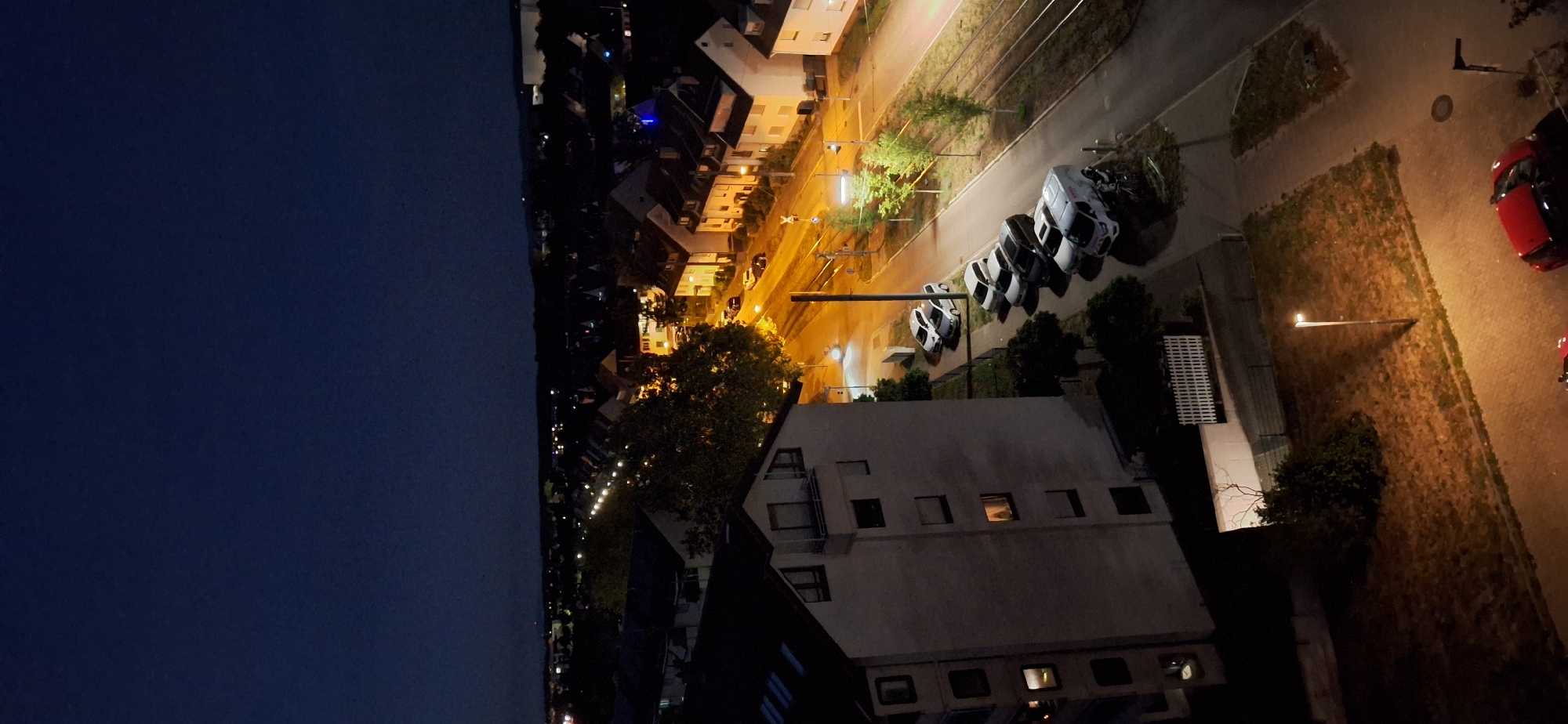 An aerial, high-angle, full shot shows a residential street scene at night in Karlsruhe, Germany. The sky is dark blue, and a few distant lights are visible on the horizon. The foreground is dominated by a multi-story building on the left, with several windows lit from within. A white picket fence and some bushes are visible in front of the building.
The street runs diagonally from the top right to the bottom left. On the right side of the street, there are several parked cars, including a white van. A bicycle is parked next to the van. Streetlights illuminate the street and the parked cars. Further down the street, more houses and trees are visible, with lights on in some of the houses. A set of tram tracks runs along the right side of the street.
In the bottom right corner, a red car is parked on a brick-paved area. A lamppost stands in the middle of a patch of dry grass. The overall impression is one of a quiet, residential neighborhood at night.