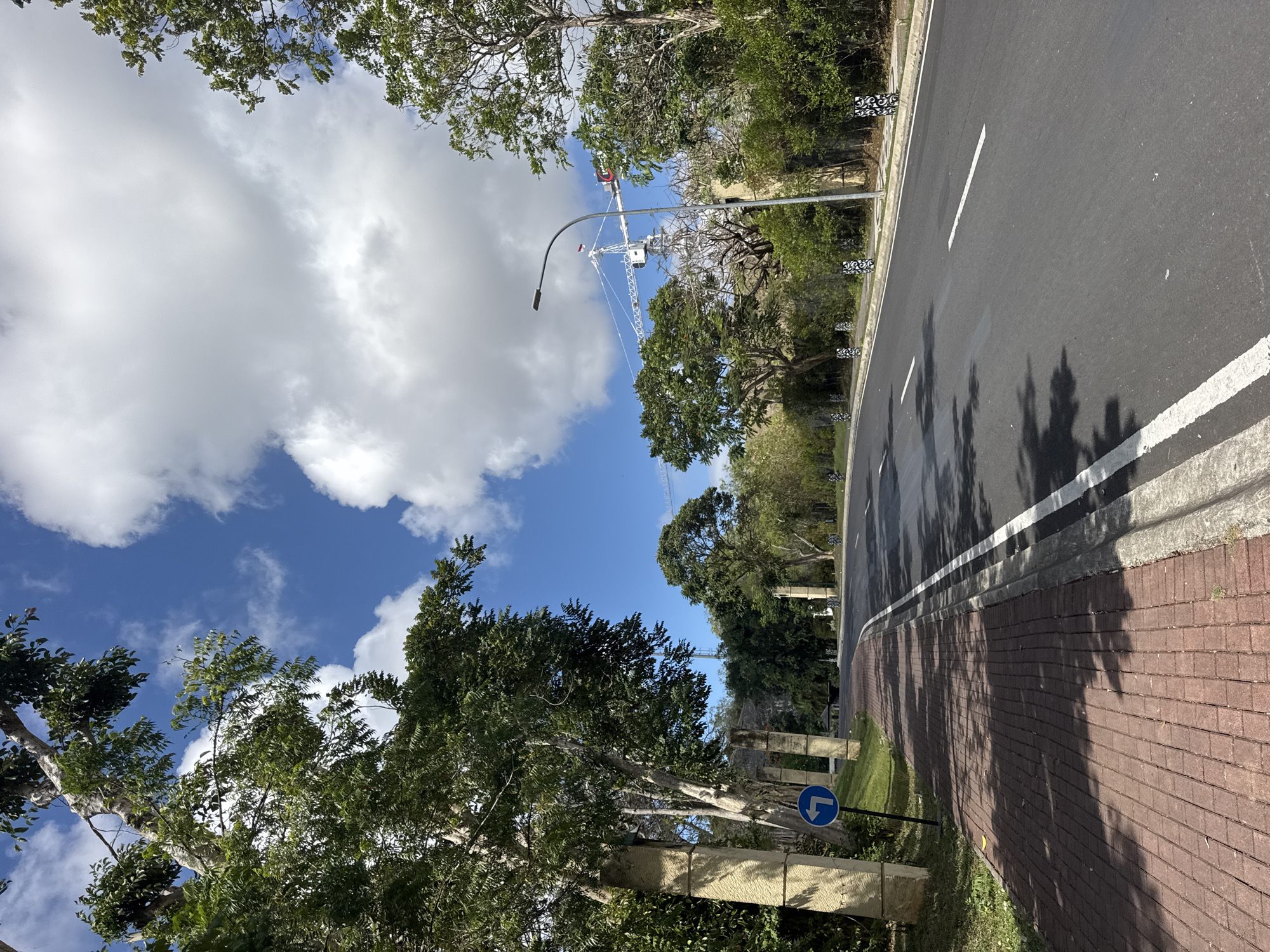 The image depicts a paved road curving to the right, flanked by a red brick sidewalk on the left and lush green trees and vegetation on the right. A tall, modern streetlamp stands on the right side of the road. In the distance, a large construction crane is visible against a bright blue sky with scattered white clouds. Several large, square stone pillars are present along the sidewalk on the left, with one featuring a blue circular sign indicating a left turn. The scene appears to be an entrance road to a development or resort in a tropical or subtropical climate, likely in Kuta Selatan, Indonesia, given the presence of tropical foliage and the potential for construction activity in developing tourist areas. There are no people or animals visible in the image. The overall impression is one of a well-maintained, modern infrastructure surrounded by natural greenery under a clear, sunny sky.