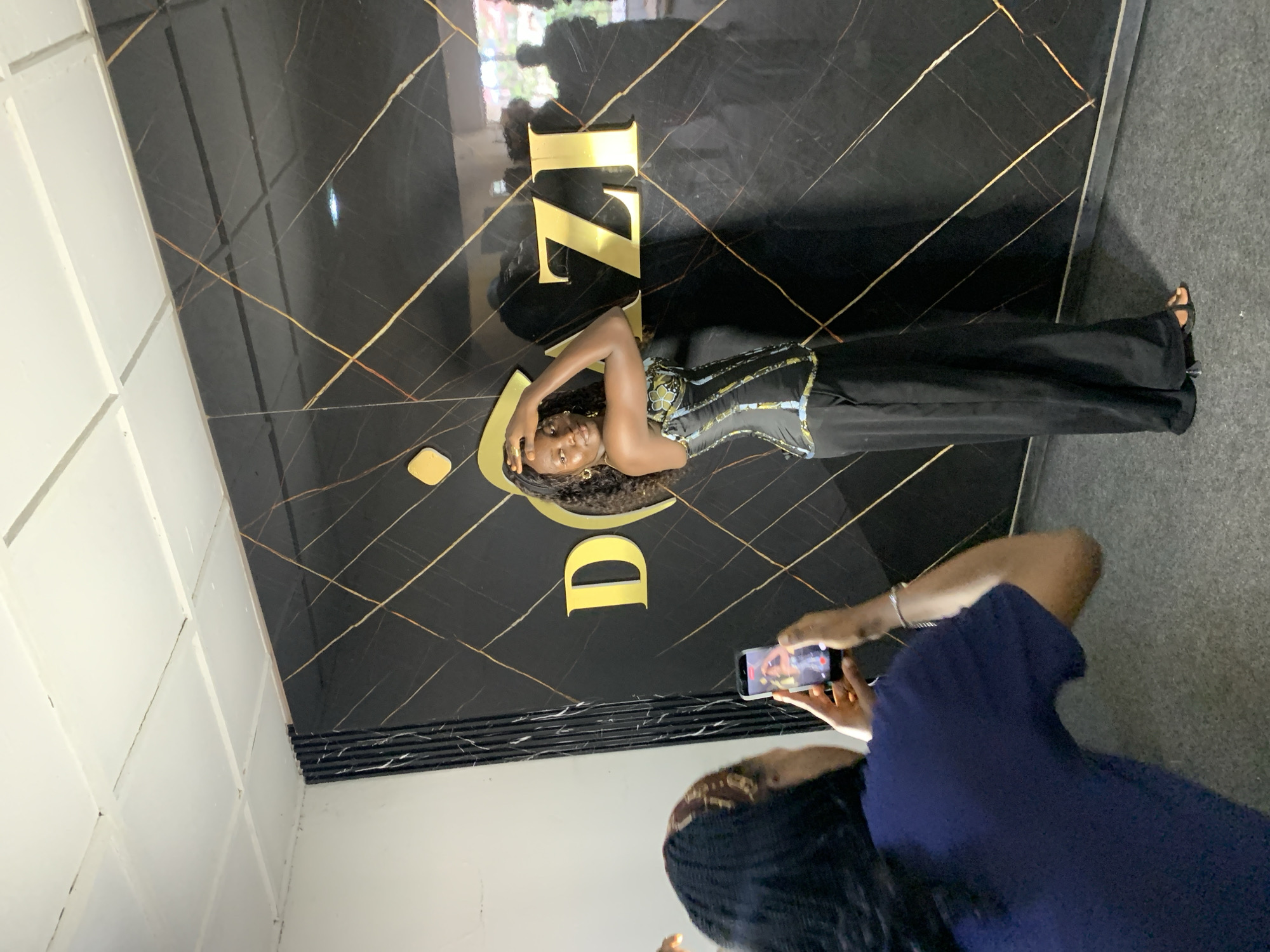 A woman is posing for a photo in front of a black marble wall with gold accents. She is wearing a black corset with gold and blue embellishments, and black flared pants. Her hair is long and dark, and she is wearing gold earrings. A person is standing in front of her, taking a picture with a smartphone. The scene appears to be indoors, with white tiled walls on the left side of the image and a grey floor. The lighting suggests it could be daytime. The text visible on the wall appears to be "TZ" and "D", possibly part of a brand name or logo. The overall atmosphere suggests a fashion or modeling shoot.