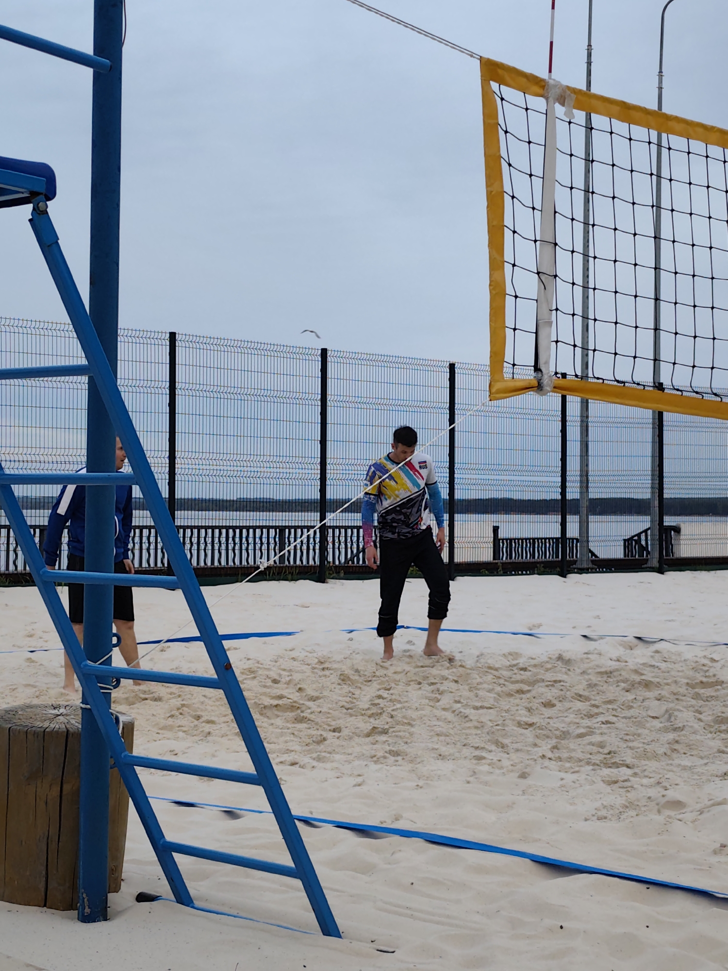 The image depicts a beach volleyball court in Chaikovsky, Russia. Two men are present; one is visible from the side near a blue volleyball post, wearing a blue jacket and black shorts. The second man stands barefoot in the sand near the net, wearing a colorful long-sleeved jersey with "RUS" visible on the shoulder, and black pants. A volleyball net with a yellow border divides the court. The ground is sandy, marked with blue boundary lines. In the background is a metal fence, beyond which lies a body of water and a wooded area. The sky is overcast.