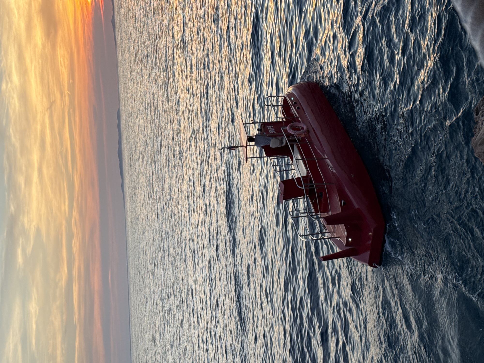 A small red submersible with a canopy is sailing on the sea near the coast of Primošten, Croatia. The submersible has a single occupant at the helm, facing away from the camera. The sun is setting, casting a warm glow over the sky and reflecting on the water. The sea is calm with gentle ripples. In the distance, faint outlines of islands can be seen. The submersible appears to be moving from left to right in the frame. The scene evokes a sense of tranquility and natural beauty.
