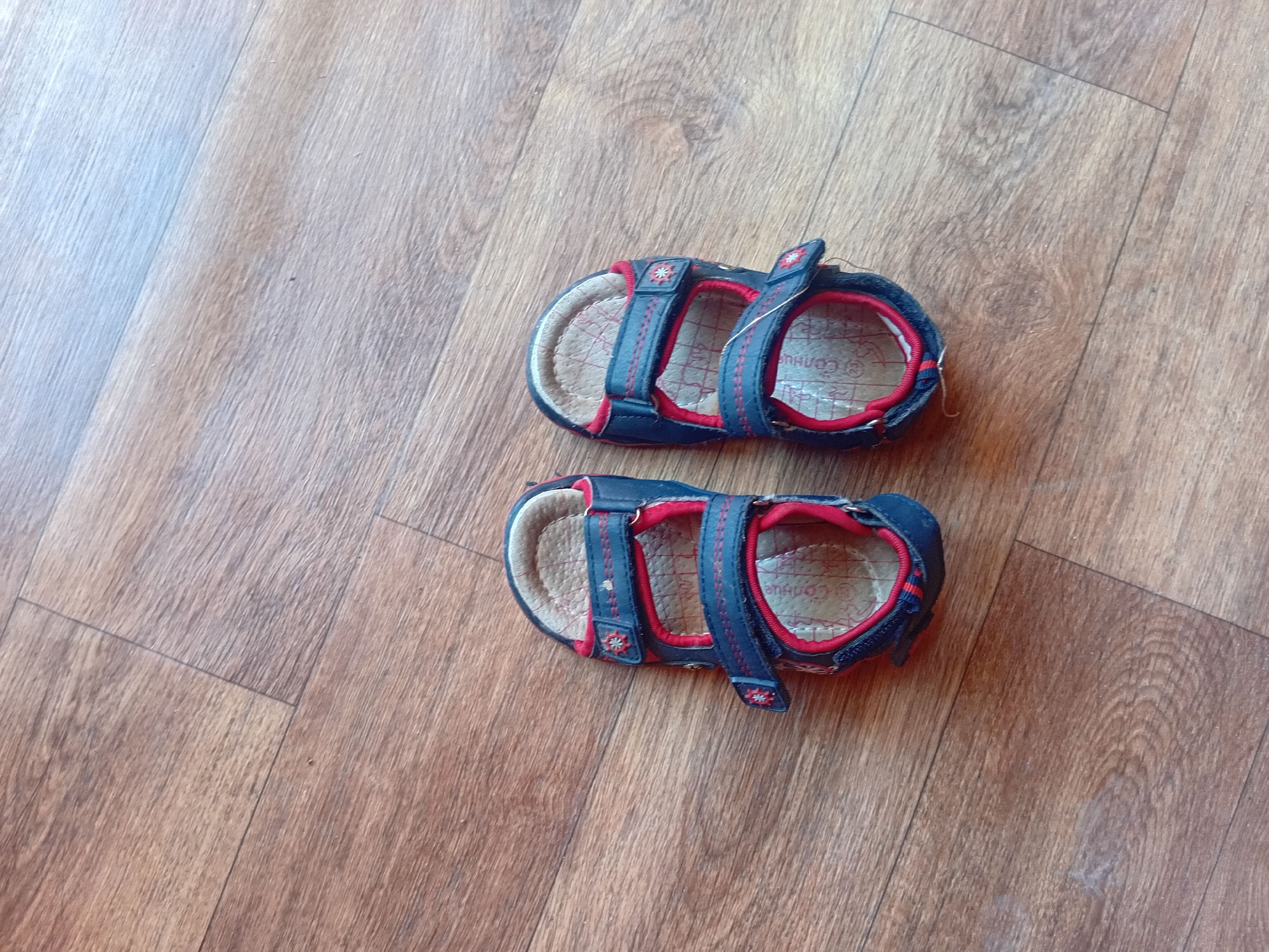 Two blue and red children's sandals are lying on a wooden floor. The sandals have velcro straps and a tan sole. The floor is made of planks of wood with a visible grain pattern. The scene appears to be indoors, in what could be a home or a storage area. There is no indication of the location being in the city of Verkhniachka, Ukraine.