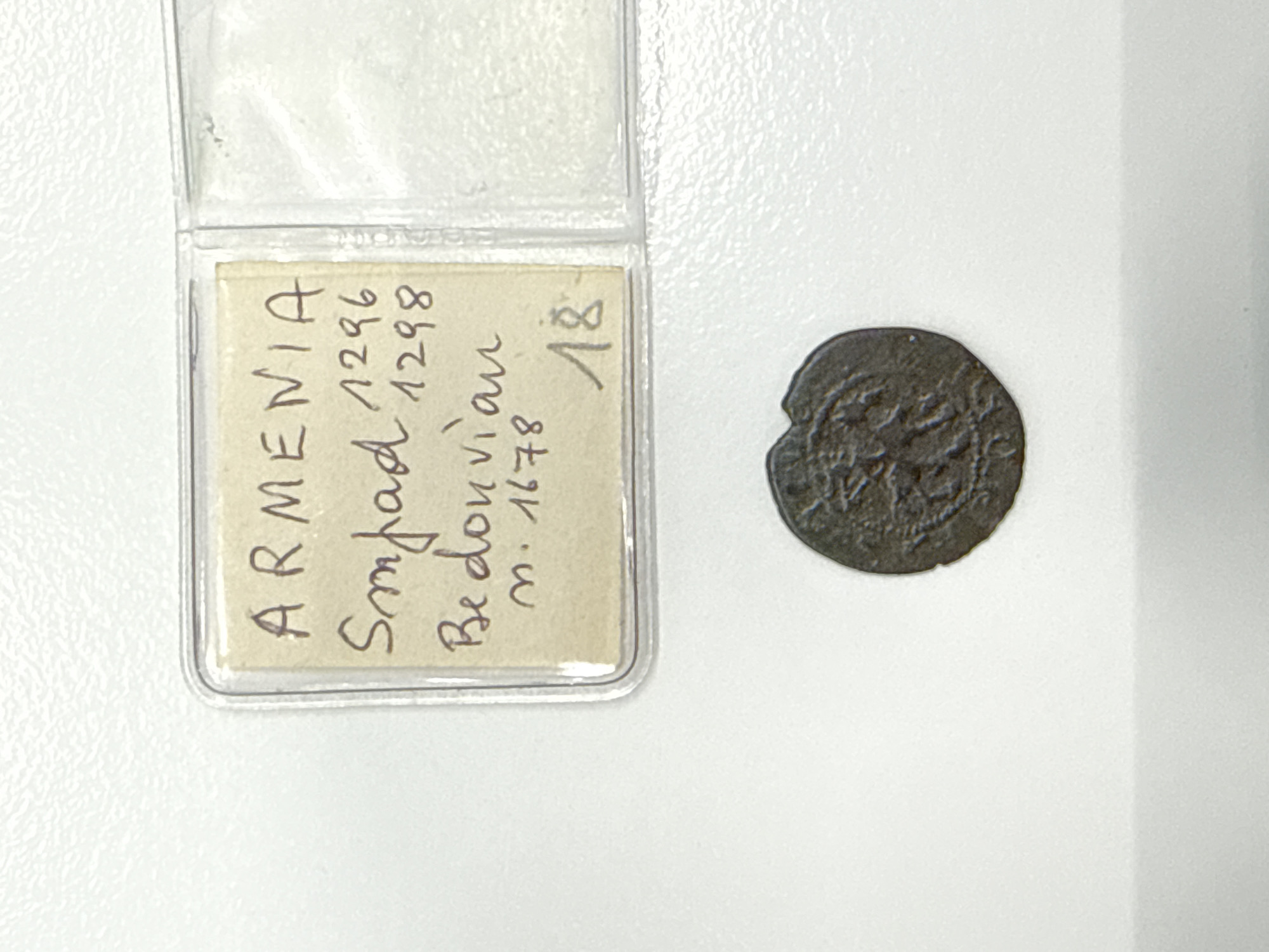 The image displays a coin and a piece of paper with handwritten text, enclosed within a plastic protective case. The text on the paper reads "ARMENIA", "Smbad 1296", "1298", "Bedouian", "m. 1678", and "18". Below the plastic case, a single coin is visible, appearing to be made of a dark metal and featuring some embossed design. The background is a plain, white, textured surface, likely a table or desk. No individuals or specific locations within Milan, Italy are depicted in the image.