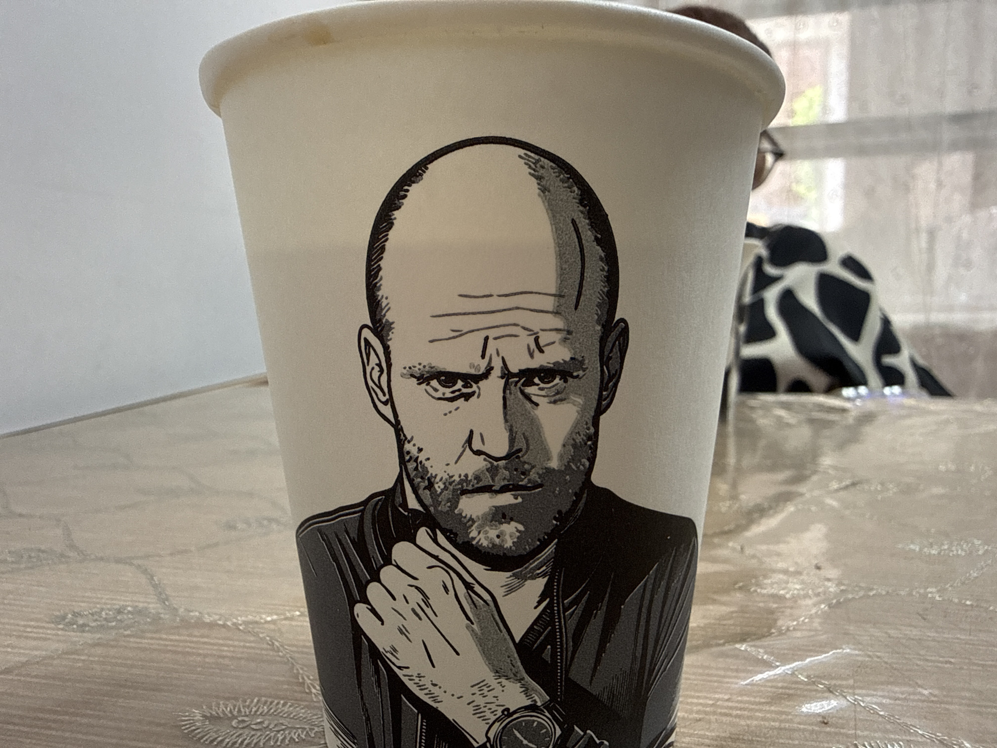 A close-up, eye-level shot shows a disposable paper cup with a black and white illustration of a man's face printed on it. The man has a bald head, furrowed brow, beard, and is wearing a dark jacket. His left hand is raised and resting on his chin. The cup sits on a light-colored, patterned tablecloth. In the background, a blurred person wearing a black and white patterned garment is visible behind a sheer curtain. The lighting suggests an indoor setting, possibly a home or cafe. The context of Beslan, Russia is not visually evident in this image.