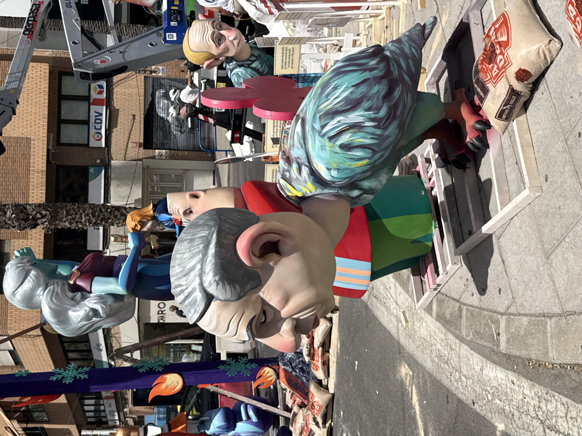This media file shows a large, colorful sculpture of human figures and a bird-like creature. The sculpture is made of wood and other materials, and it is painted in bright, bold colors. The figures are depicted in various poses, and they appear to be engaged in some kind of activity or celebration. The sculpture is situated outdoors, in what appears to be a public space with buildings and a paved ground. A crane is visible in the background, suggesting that the sculpture may be part of a larger construction or installation project. There are also signs with text in Spanish, including "gerpasa", "CDV", "OIL", "CHARO", "ADENAS DE", and "ARENAS EL VALLE". The overall impression is one of a vibrant and festive event, possibly a parade or festival. The lighting suggests it is daytime.