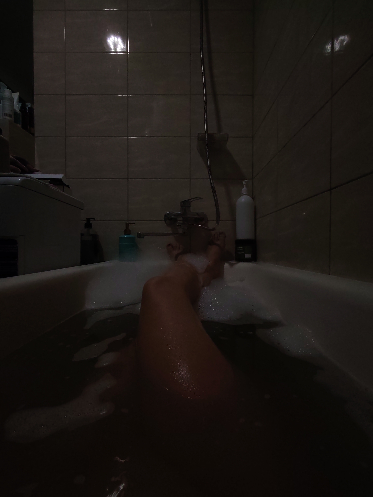 A low-angle, dimly lit indoor shot shows a person's legs submerged in a bubble-filled bathtub. The bathtub is white with a metallic faucet. On the edge of the tub, to the left, are two pump-action soap dispensers, one black and one light blue. Further left is a white washing machine with a dark object on top. The wall behind the tub is tiled with square, light brown tiles. A shower head and hose hang on the wall to the right. The image appears to be taken in a residential bathroom. There are no identifiable landmarks or contextual clues to indicate the location in Izhevsk, Russia.