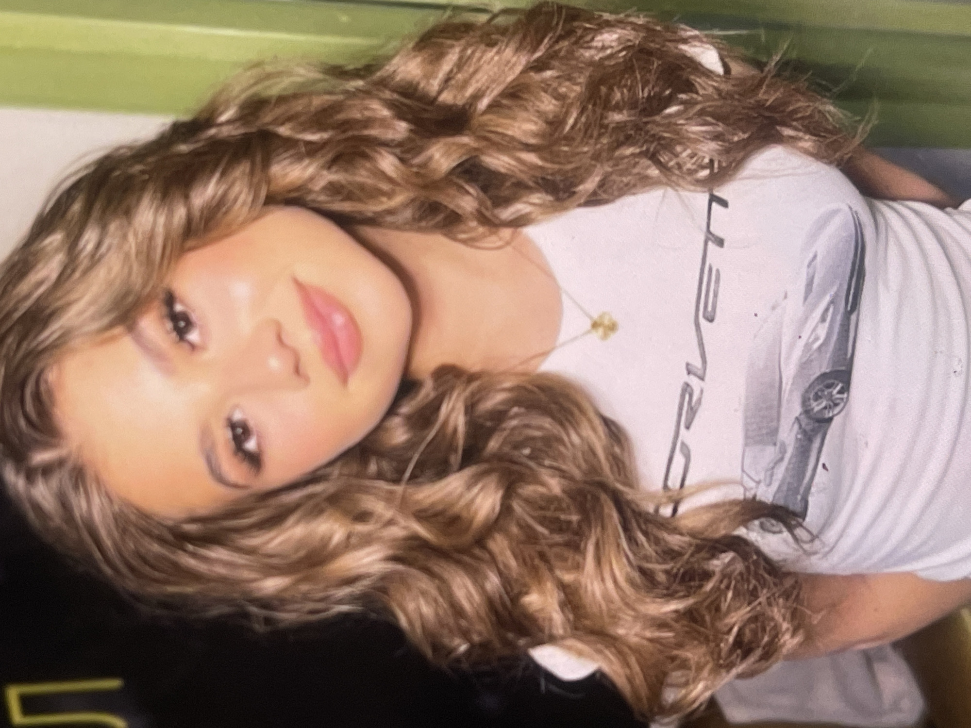 A close-up portrait features a young woman with long, voluminous, light brown hair styled in soft, cascading curls and waves, accentuated with lighter highlights. Her head is slightly tilted, and she gazes directly at the viewer with a serene expression and a hint of a smile on her pink-tinted lips. She wears natural-looking makeup, including defined eyebrows, subtle eyeliner, mascara, and blush.
She is dressed in a white crew-neck t-shirt. A prominent graphic on the shirt depicts a sleek, light gray sports car, viewed from the side-front, likely a Corvette. Vertically aligned text, partially visible next to the car, clearly spells "CORVETTE". A delicate gold-toned necklace with a small, intricate charm is visible around her neck.
The background is softly blurred, showing an upper section of light green and a lower section of a dark, possibly black or dark green, surface, indicating an indoor setting. The overall impression is one of casual elegance and a calm demeanor. This image was taken in Poltava, Ukraine.