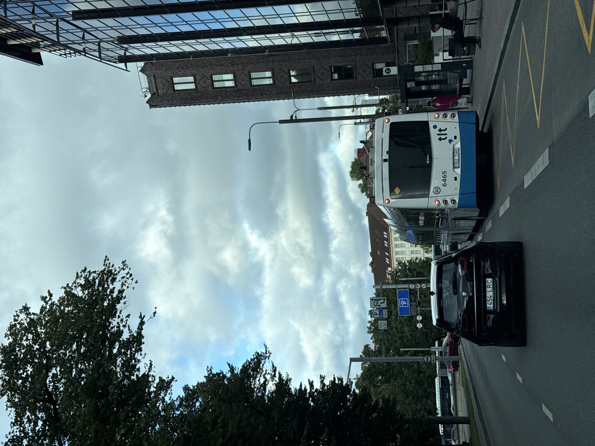 A street view in Tallinn, Estonia, shows a black BMW i3 with license plate "454 LRC" driving on the right side of the road. A white and blue trolleybus, number 6465, with the logo "tlt" is in front of the BMW. The trolleybus is stopped at a bus stop where two people are waiting. A modern glass-fronted building with dark brick accents is on the right side of the street. A brick building is visible behind the glass building. Trees line the left side of the road, with a park visible in the background. Traffic signs indicating parking regulations are present. The sky is cloudy.