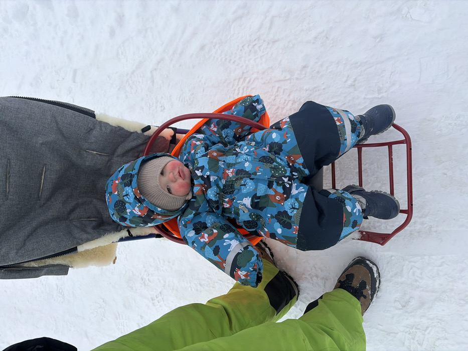 A baby or toddler, dressed in a blue patterned winter jumpsuit depicting forest animals, a beige knit hat, and blue winter boots, sits in a red metal sled with an orange backing. The child's cheeks exhibit a rosy coloration. To the left, the lower legs and feet of an adult are visible, clad in lime green snow pants and brown winter boots. Behind the child and to the right, a gray fabric child stroller or carriage, featuring a cream-colored sheepskin lining, is partially visible. The scene is set outdoors on a uniformly snow-covered ground in the city of Chaikovsky, Russia.