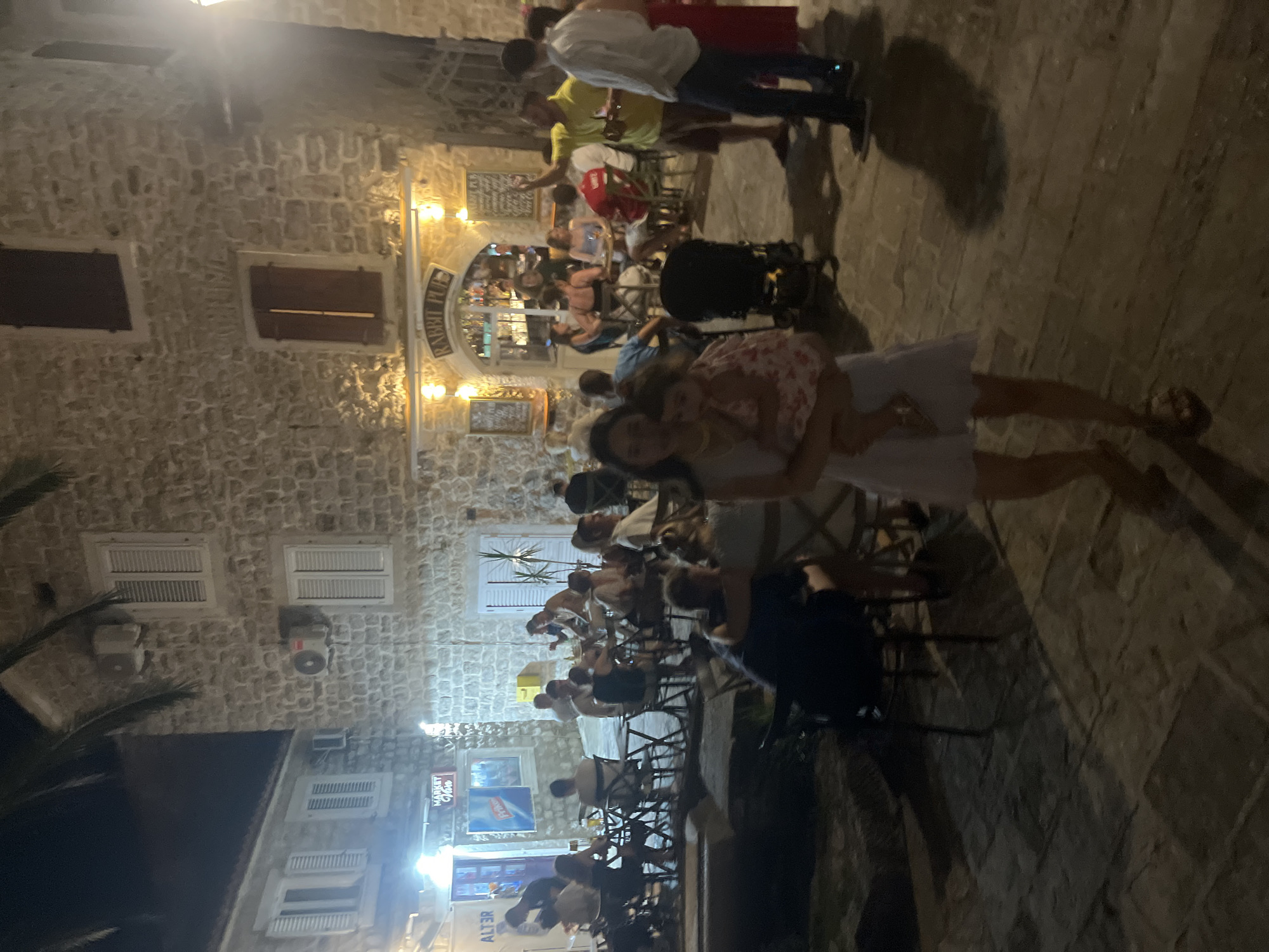 The image captures an evening scene in Budva, Montenegro, likely on a cobbled street. Several people are gathered outside what appears to be a restaurant or pub, as indicated by tables and chairs arranged on the pavement. The lighting suggests it is nighttime, with artificial lights illuminating the stone building facade and the street.
The subjects include a mix of adults and children. In the foreground, a woman is holding a young child, both looking towards the camera. In the background, groups of people are seated at tables, engaged in conversation and enjoying their evening. Some are eating or drinking. The architecture is characteristic of old European towns, with stone walls and wooden shutters on the windows.
The atmosphere seems relaxed and social, with people enjoying the warm evening. There are some decorative elements visible, including lights and signage. One sign above an arched entrance reads "RABBIT PUB." Another sign visible on a building in the background says "MARKET VIDEO." The overall impression is that of a lively evening at an outdoor dining establishment.