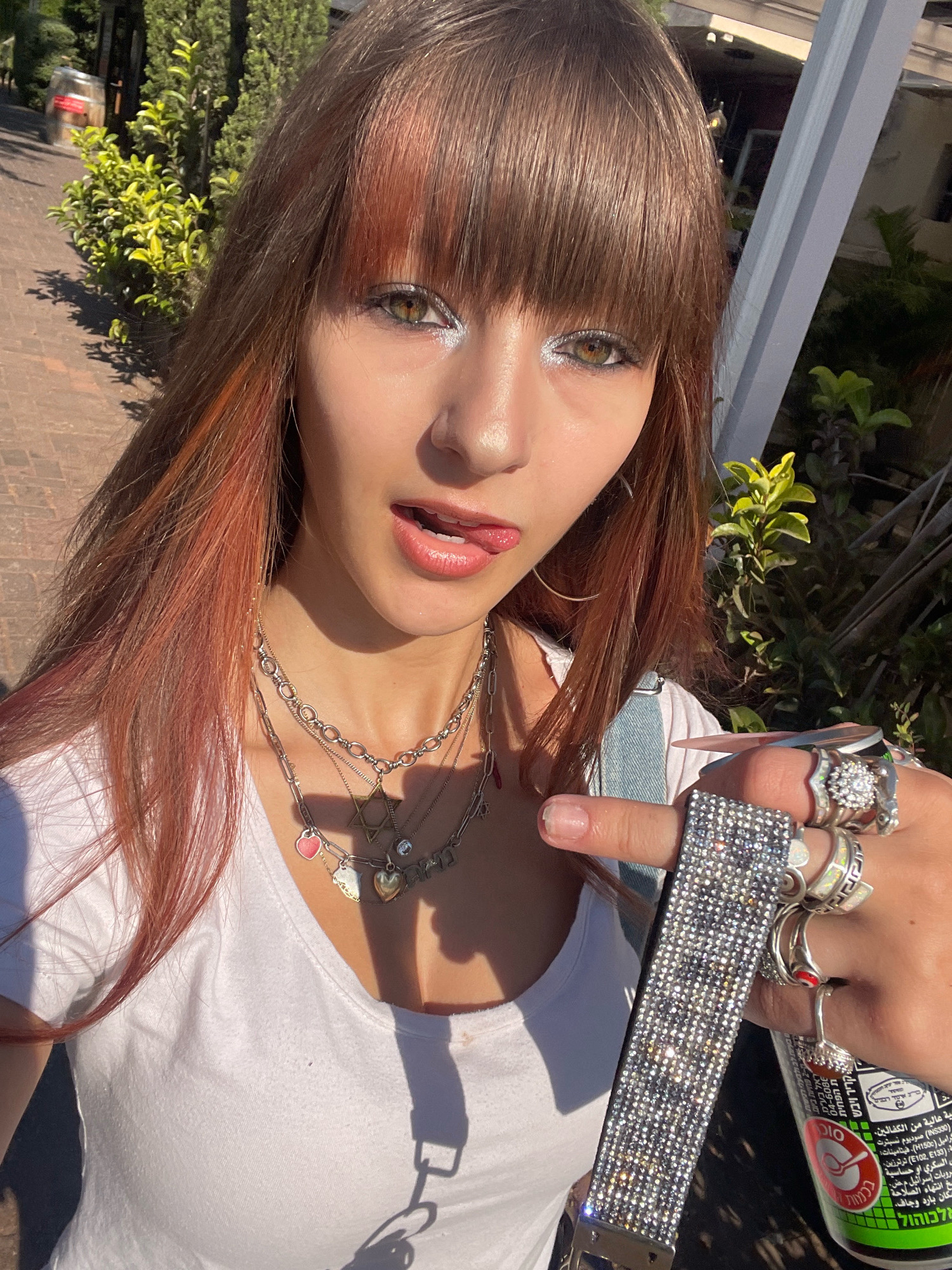 A young woman with long brown hair with copper highlights and bangs is shown from the chest up, sticking her tongue out playfully. She is wearing a white t-shirt and multiple silver necklaces with charms. She is also wearing several large, ornate silver rings on her fingers, and is holding a sparkling, studded bracelet. Behind her, green bushes and trees are visible, as well as a brick walkway. The lighting suggests it is daytime. The location is Raanana, Israel.