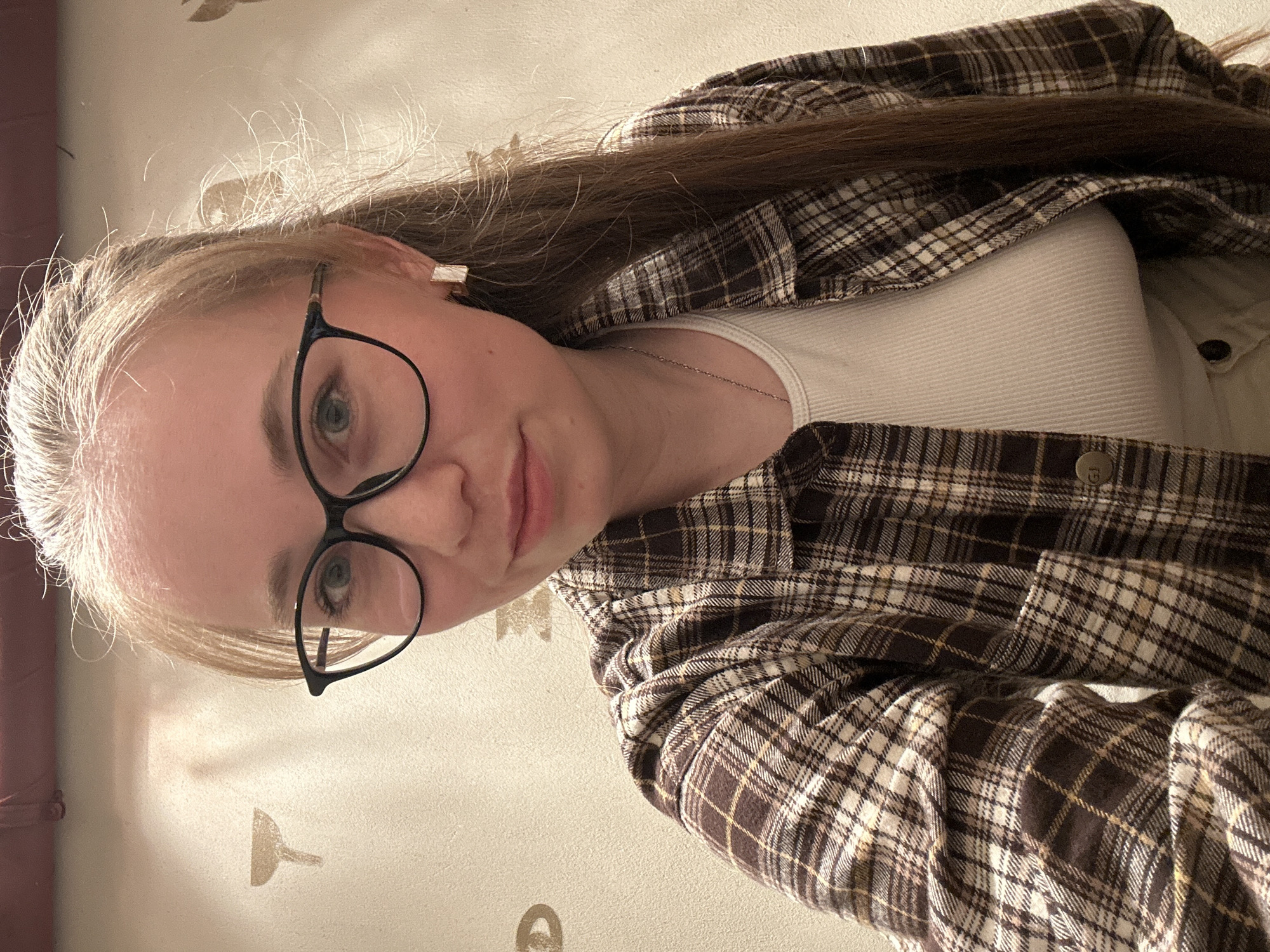 A young woman with fair skin and light blonde hair, partially tied back, is wearing black-rimmed glasses. She is wearing a white tank top under a brown and tan plaid flannel shirt. A silver necklace is visible around her neck. She is indoors. The image orientation is rotated. The background is a light beige wall with faint decorative patterns. The location is not identifiable as Saint Petersburg, Russia from the image.