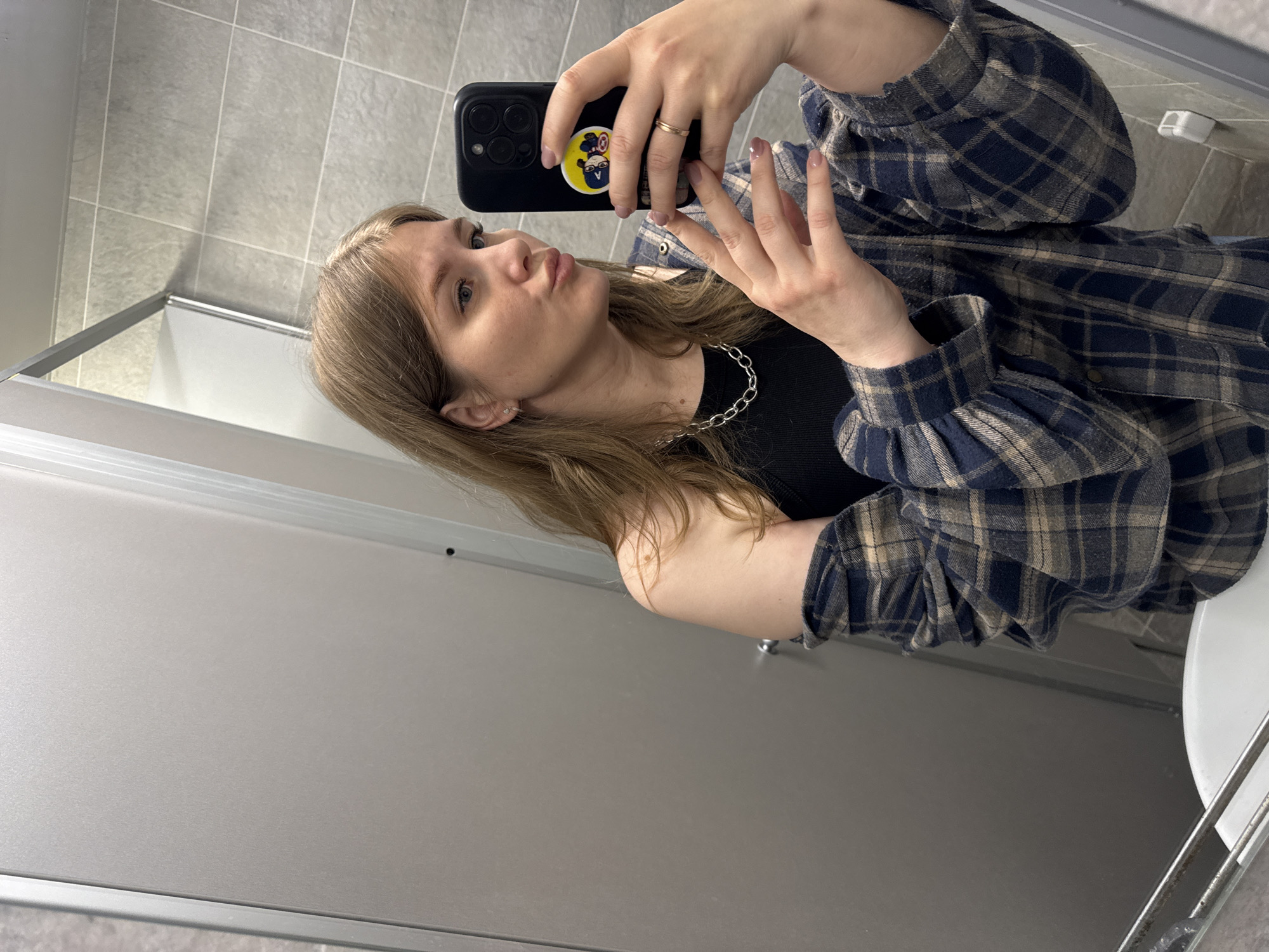 A young woman is taking a selfie in what appears to be a public restroom. She is holding a black smartphone with a Captain America sticker on it. She has fair skin, light brown hair, and is wearing a black top with a silver chain necklace and a plaid shirt. She has a ring on her finger. The background consists of gray tile walls, a bathroom stall, and a white sink. The location could be in Perm, Russia, but there is no specific evidence in the image to confirm this.