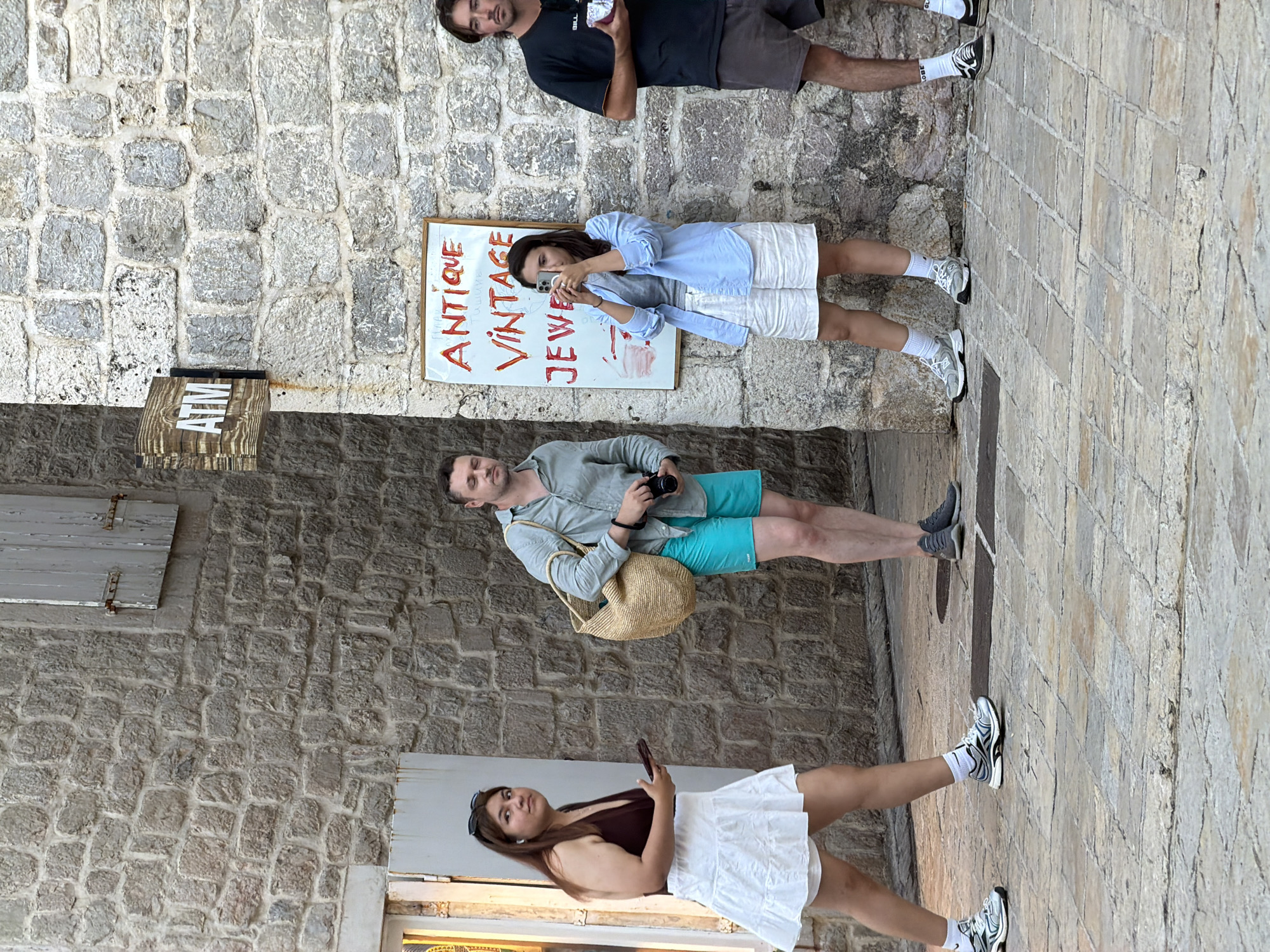 The image depicts an outdoor scene on a cobblestone street in the Old Town of Budva, Montenegro. A group of four people are present. On the left, a woman with long brown hair wears a dark sleeveless top and a white skirt, holding a phone in her right hand and looking away from the camera. Next to her, a man in a light blue button-down shirt and turquoise shorts is holding a camera and a woven bag. In the center, a woman in a light blue shirt and white shorts is taking a photo with her phone. To the right, a man in a black t-shirt and grey shorts stands with a white object in his hands. Behind the people, a stone wall is visible with a sign for an ATM and another sign that reads "ANTIQUE VINTAGE JEWELRY" in red lettering. The architecture suggests a historic urban setting.