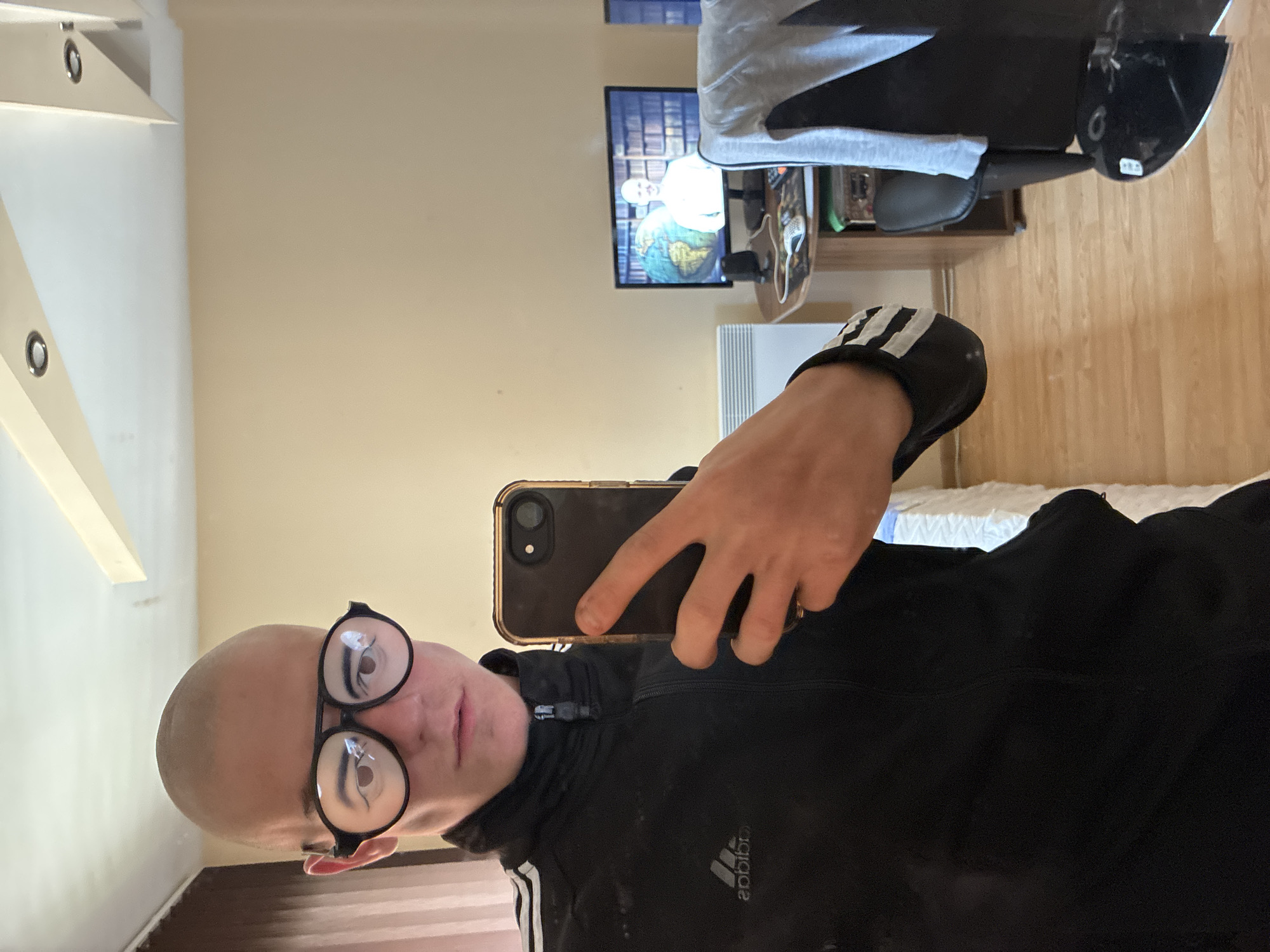 A person is seen taking a selfie in an indoor setting, likely a room in Varna, Bulgaria. The image is taken from a first-person perspective, with the person holding a black smartphone to capture their reflection.
The subject is a light-skinned individual, appearing male, with a bald or very closely shaved head. They wear large, round, black-framed glasses and a black Adidas full-zip jacket with white stripes on the sleeves. Their expression is neutral.
The background reveals a cozy room with light beige walls and a light wooden floor. A white ceiling features decorative or structural elements. On the right side of the frame, a bed with white bedding is partially visible. Further back, an office chair with a light grey towel draped over it is positioned near a wooden shelf or desk. Above the desk, a flat-screen television is mounted on the wall, displaying a scene with a person, a large globe, and bookshelves. A white radiator is visible on the wall beneath the TV.
Visible text includes the "adidas" logo on the jacket (reversed due to the reflection), and indistinct text on a tag beneath the chair. The time of day is not clearly indicated, suggesting indoor lighting conditions.