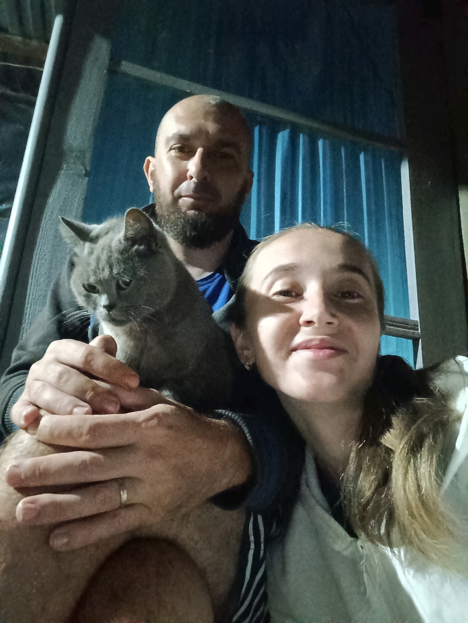 A bald, bearded man holds a gray cat while a woman with long, blonde hair smiles at the camera. The man is wearing a dark hoodie over a blue t-shirt. The woman is wearing a white hoodie. They are in an outdoor setting, possibly a porch or balcony, with a blue corrugated metal wall in the background. A metal railing is visible on the left. The image is dark, suggesting it was taken at night or in low light conditions. The woman's face is illuminated by a light source, creating a shadow on the left side of her face. The man's left hand, wearing a wedding ring, rests on the cat.