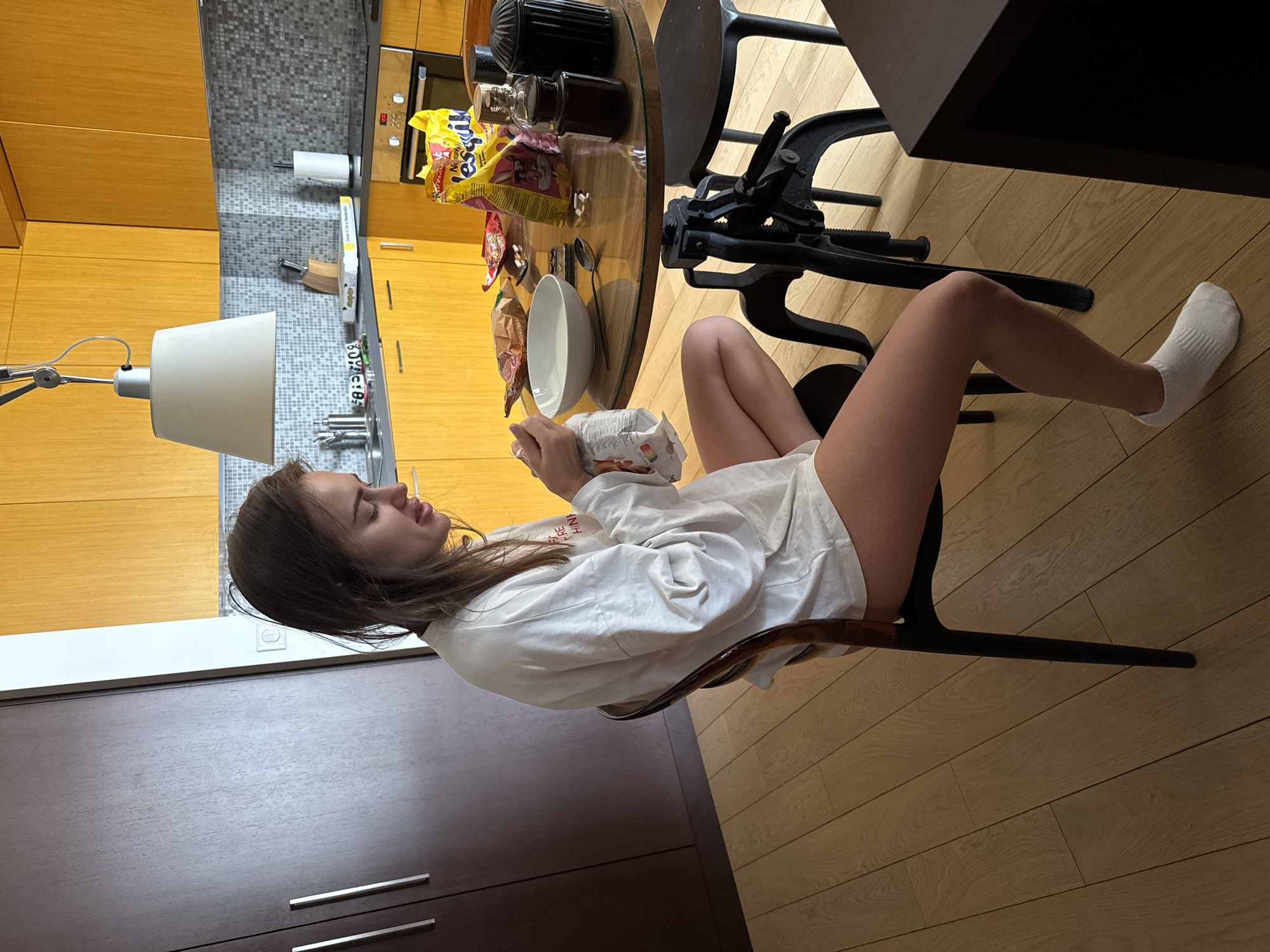 A young woman sits on a chair at a table in an apartment kitchen in Warsaw, Poland. She wears a white oversized shirt and a white sock on her right foot. She is holding and eating from a white and patterned snack package. The table holds a white bowl, snacks, and jars. The kitchen features brown and yellow cabinets, a tiled backsplash, and a lamp over the counter. The floor is light-colored hardwood.