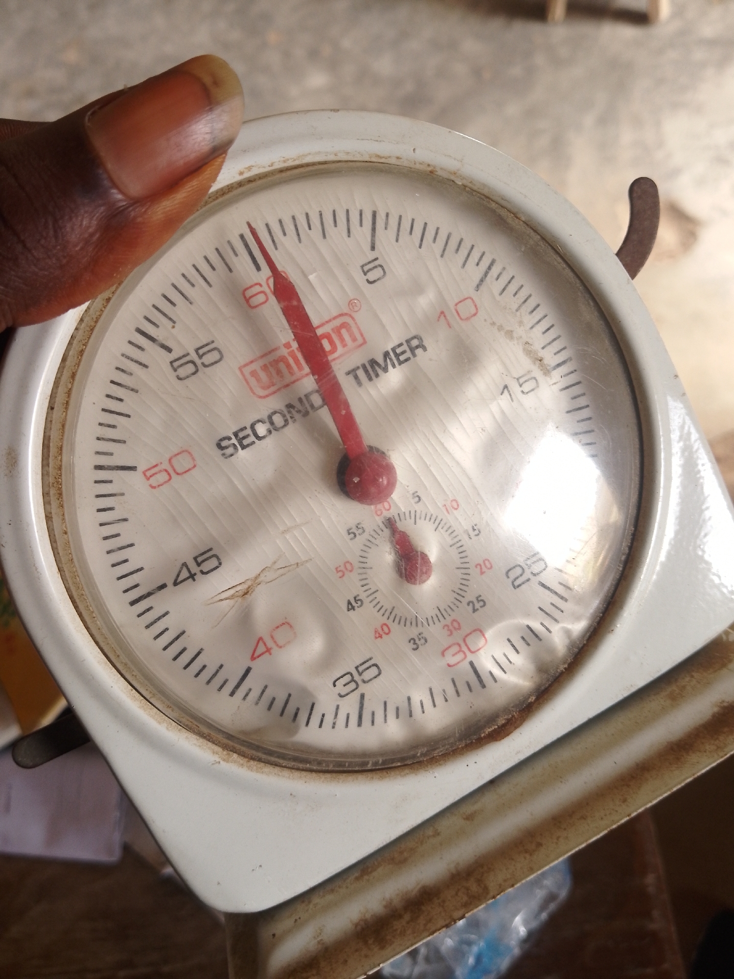 A close-up, top-down shot captures a person's thumb and index finger holding an analog stopwatch. The stopwatch, predominantly white with some rust-colored stains, displays a circular dial with black minute markings and red numerical labels from 0 to 60. A red hand points to the 60-minute mark, indicating the stopwatch is at its starting position. A smaller dial within the main dial shows finer time increments. The stopwatch is part of a larger, white object that appears to be a scale, with its edge visible at the bottom of the frame. The background is out of focus, showing a textured, light-colored surface that could be a floor or ground. The lighting is natural and appears to be from overhead.