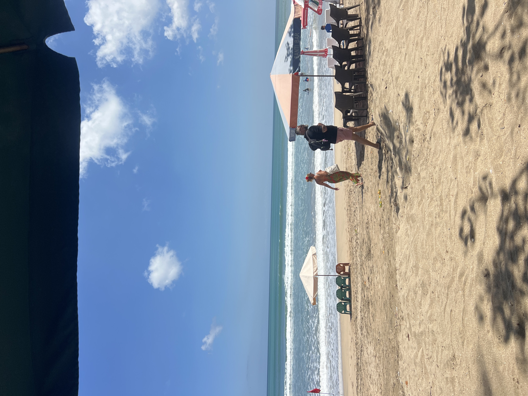 On a sunny day at Kuta Beach, Indonesia, the ocean meets a sandy shore. The turquoise waves are gentle, with white foam cresting as they approach the beach. A few people are visible in the water, enjoying the ocean. Along the shore, beach chairs and umbrellas are set up, some occupied and some empty, indicating a public beach area.
In the foreground, the sand is warm and textured, with shadows of palm leaves cast across it from overhead foliage, suggesting a shaded area. Two individuals are walking on the beach. A woman in a patterned dress and a woman with sunglasses are walking away from the camera towards the ocean. Behind them, a man wearing a black shirt and pink shorts walks with a backpack.
Further down the beach, more umbrellas and seating areas are visible, with patrons seated at tables under one of the larger umbrellas. A red flag is planted in the sand near the water's edge, possibly indicating a swimming zone or safety warning. The sky is a vibrant blue, dotted with fluffy white clouds, contributing to the idyllic beach scene. The overall impression is one of a relaxed and pleasant day at a popular tourist destination.