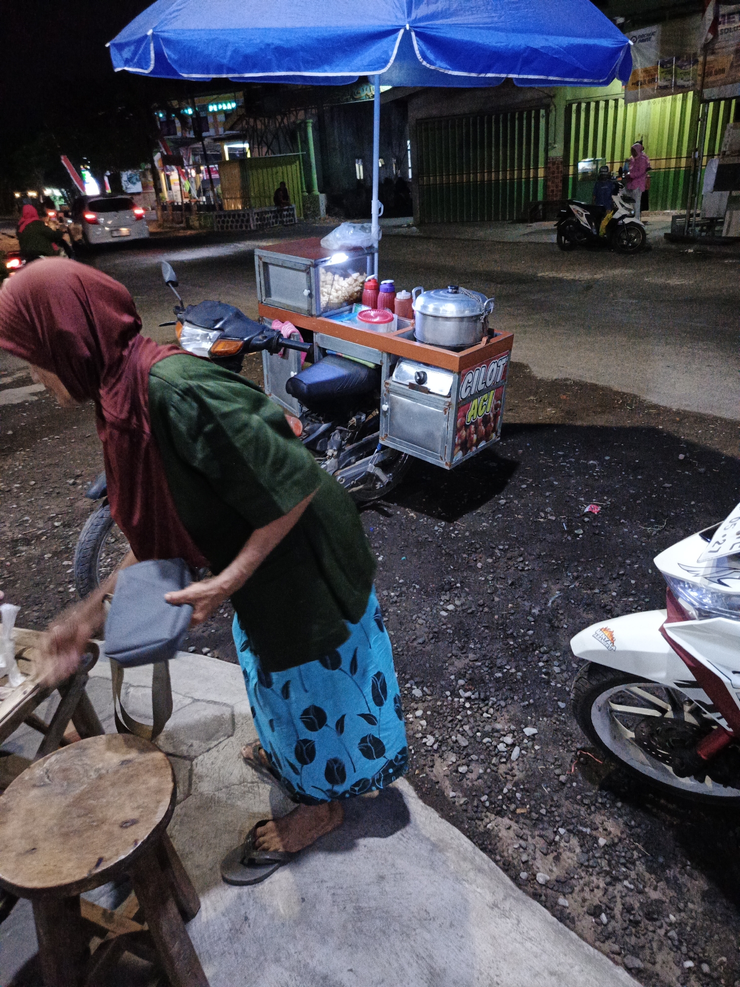 A woman wearing a maroon headscarf and a dark green shirt, and a blue skirt with a black floral pattern, stands near a mobile food cart. She is holding a gray bag in her right hand. The food cart, which is mounted on a motorcycle, has a blue umbrella canopy and displays the text "CILOT ACA" on its side, along with images of food items. There are several containers of food and sauces on the cart. To the left of the woman is a small, round wooden stool. The scene takes place at night on a street in Sumbersari, Indonesia, with other vehicles and buildings visible in the background.