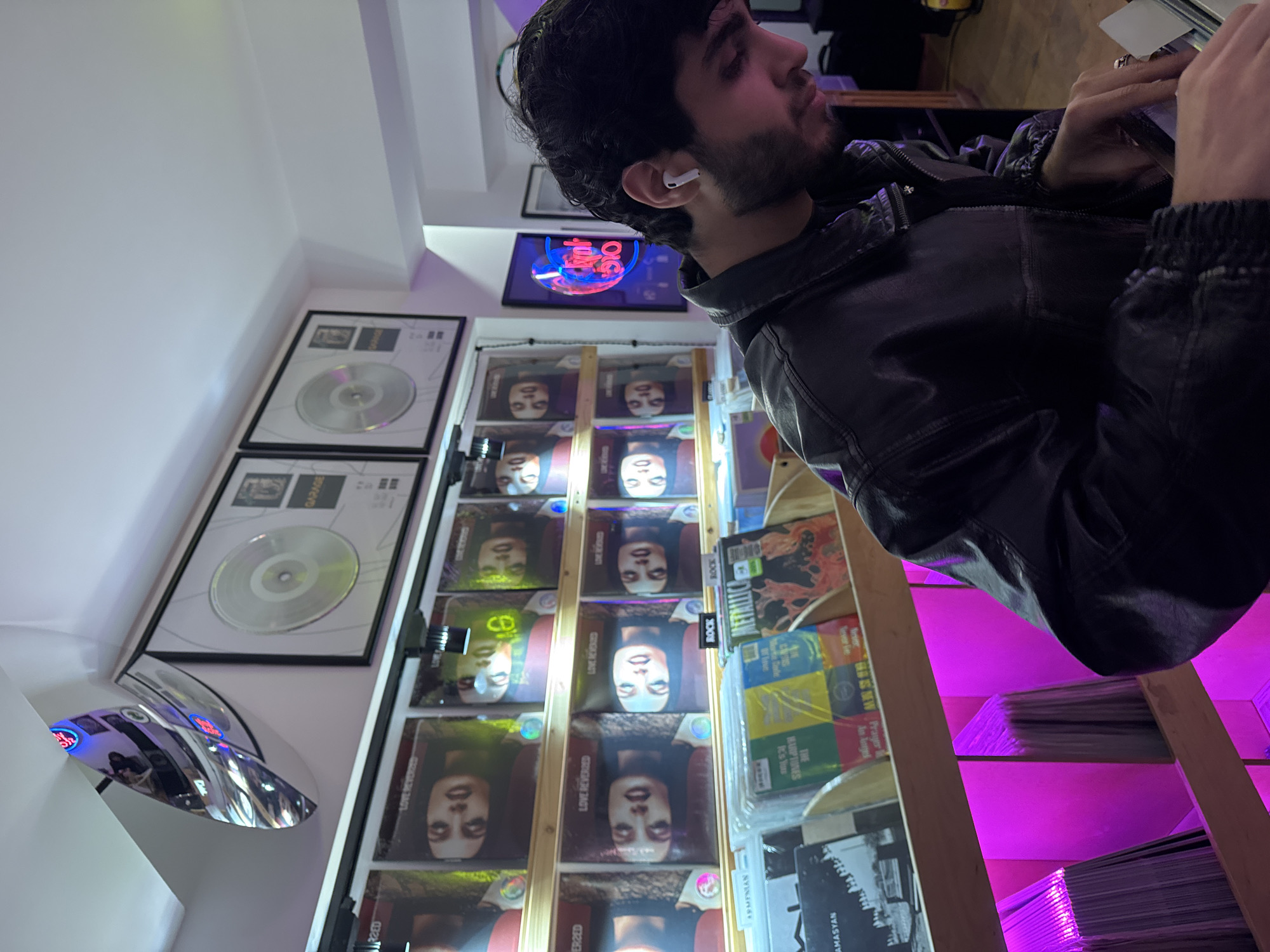 A man with dark hair and a beard, wearing a black leather jacket and an earbud in his left ear, is pictured in what appears to be a record store. Behind him are shelves displaying rows of vinyl records, each with the same cover image. Above these records are framed platinum records mounted on the wall. Additionally, there is a neon sign that says "40 OLG". Additional records can be seen stacked on the shelves. The setting suggests a modern, urban environment, possibly within the city of Yerevan, Armenia.