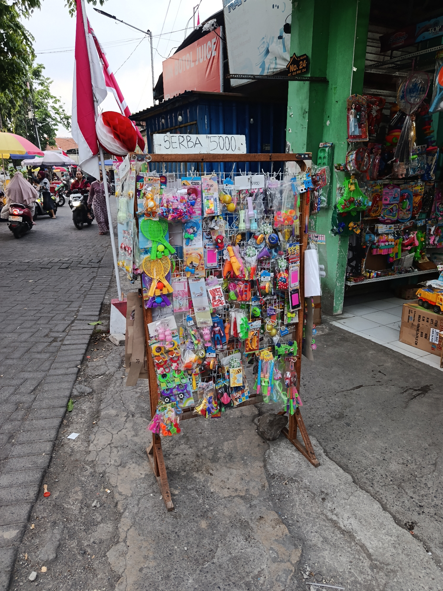 The image shows a street scene in Keboansikep, Indonesia, featuring a temporary stall selling an assortment of small toys and trinkets. A red and white Indonesian flag is prominently displayed on a pole to the left of the frame. The stall itself is a wire rack on a wooden stand, packed with colorful items like miniature rackets, plastic figures, balls, and various packaged toys. In the background, a building with green trim and a large red sign that reads "BOTOL JUICE" is visible. Several people are present, some on motorbikes, suggesting the area is a public thoroughfare. The ground is paved with cobblestones on the left and asphalt on the right, showing signs of wear and tear. Umbrellas provide shade for other stalls or vendors in the distance. The overall impression is of a busy, informal marketplace.