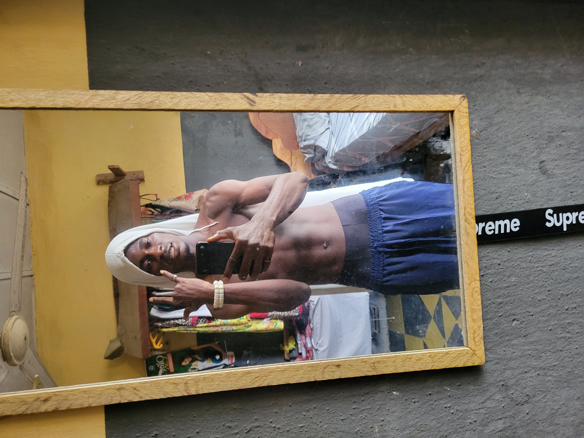 A young Black male is taking a mirror selfie in what appears to be a bedroom in Onitsha, Nigeria. He is wearing blue shorts and has a white cloth draped over his head. He is holding a black smartphone in his right hand and making a "rock on" hand gesture with his left. A wristband made of several light-colored beads is visible on his left wrist. The room is sparsely furnished, with a bed with gray bedding visible in the background, along with some clothing hanging on a rack. A portion of a white ceiling fan is visible at the top of the frame. The mirror is framed with a light-colored wood. A black strap with white text that reads "Supreme" is hanging from the bottom of the mirror.