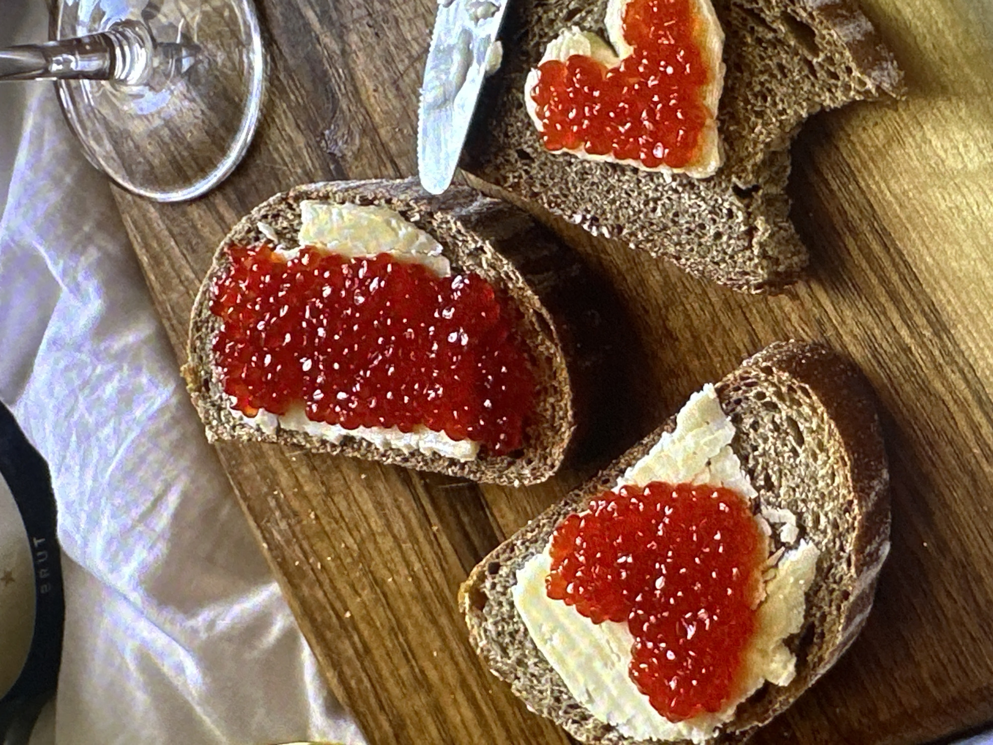 The image depicts a wooden board with three slices of dark bread topped with a layer of white spread, likely butter or cream cheese, and a generous amount of red caviar. One slice of bread is cut in a heart shape. A butter knife rests on the board between two of the bread slices. A portion of a wine glass is visible in the upper left corner, and the label of a bottle, marked "BRUT," is visible in the lower left corner. The board is set on a white wrinkled cloth. There is no evidence to suggest the location is Yerevan, Armenia.