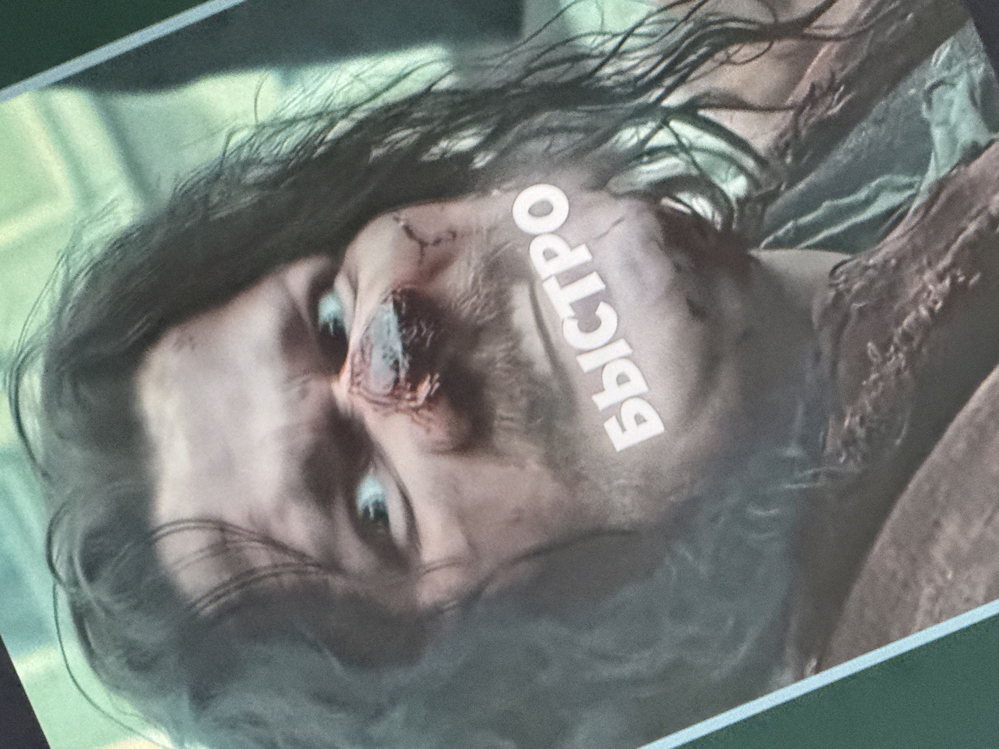 The image is a close-up, slightly blurry photograph of a man's face. He has dark, unkempt hair, a light complexion, and prominent dark circles under his eyes. His nose appears to be injured, with a dark, textured area covering the bridge and right nostril. White text, appearing to be Cyrillic letters that spell "БЫСТРО" (which translates to "FAST"), is overlaid on his lower face and neck, obscuring part of his mouth and chin. The background is out of focus and appears to be a mixture of green and brown tones, suggesting an indoor or perhaps a natural setting. There is no indication of the city of Saratov, Russia, in the image.