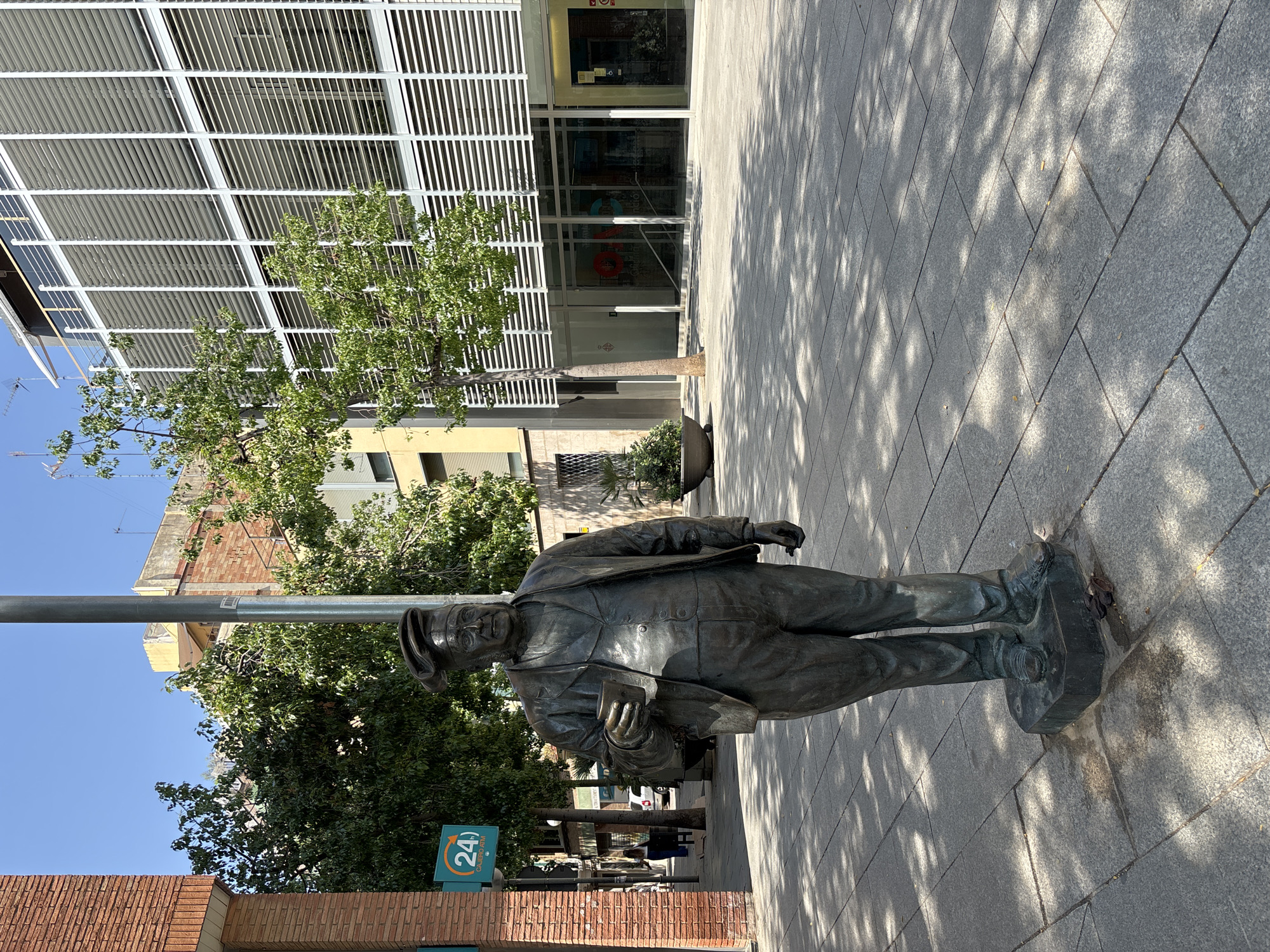 A bronze statue of a man wearing a hat, suit, and tie stands on a paved sidewalk in front of a modern building. The man is holding a small cup in his right hand. The building behind him has a facade of horizontal louvers. Trees and shadows from the trees are visible on the sidewalk. A "24h CAJERO ATM" sign is mounted on a pole to the left of the statue. In the background, there are more buildings and a few people walking. The scene appears to be in an urban setting, likely Barcelona, Spain, given the architectural style and the "24h CAJERO ATM" sign.