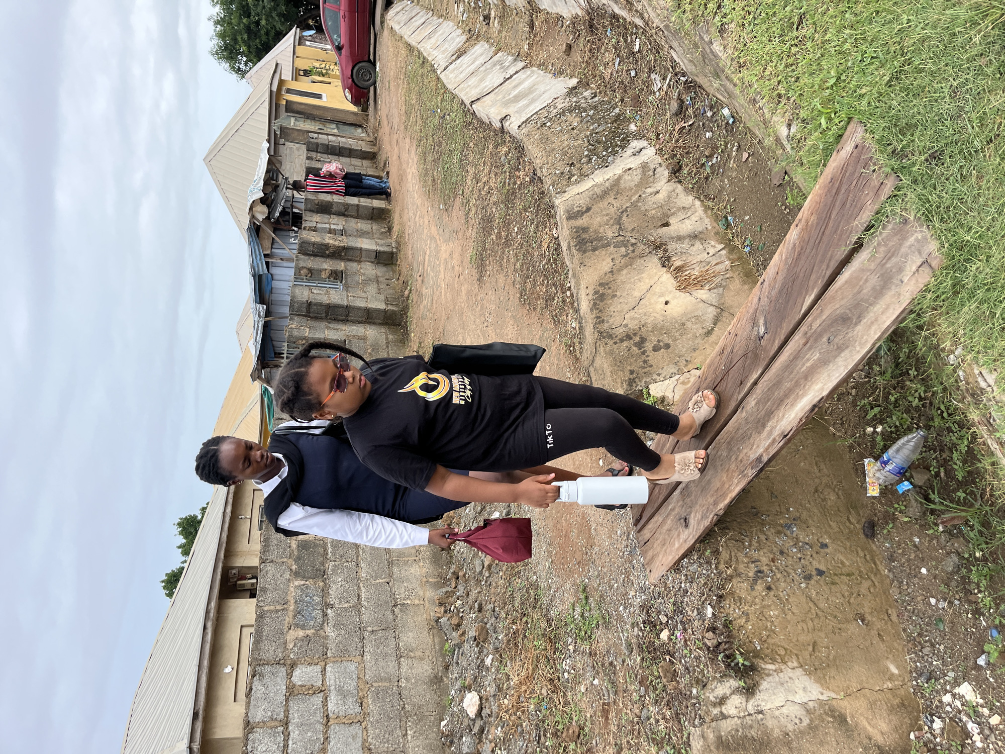 The image captures an outdoor scene, likely in a residential or semi-urban area of Abuja, Nigeria, under an overcast sky. In the foreground, two women are engaged in an interaction on an unpaved path.
The woman on the right, positioned partially on two wooden planks forming a makeshift bridge over a muddy ditch, wears a black t-shirt with a yellow and white graphic logo, black leggings with the text "T.K.T.O" visible on her left thigh, and light-colored sandals with flower embellishments. She is holding or receiving a white water bottle.
To her left, the second woman wears a white long-sleeved shirt beneath a dark vest. She holds a maroon bag in her left hand and appears to be handing the white bottle to the other woman.
The surrounding environment consists of dirt ground with sparse patches of grass and a concrete drainage channel running alongside. The ditch beneath the wooden planks contains murky water and some discarded items, including a clear plastic bottle and a white wrapper. In the mid-ground, a person wearing a red and white striped shirt and blue jeans stands near a building. The background features several simple buildings with concrete block walls and corrugated metal roofs. A red car is parked on an elevated dirt area to the upper right.