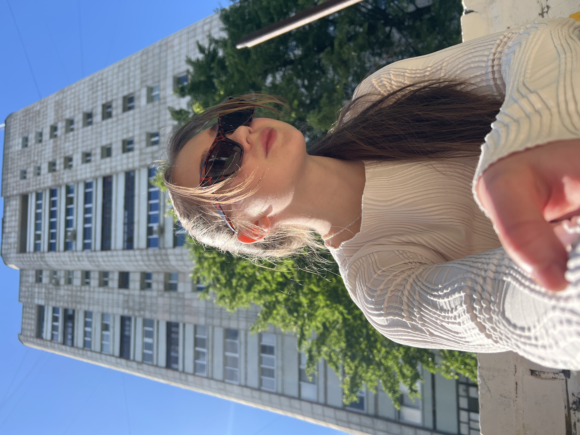 A medium shot shows a fair-skinned young woman wearing sunglasses and a light-colored, patterned top in an outdoor urban setting. She is positioned at an angle, looking upwards towards the sun. Behind her, there is a tall, Soviet-era building with a grid-like facade of windows and a tree with lush green leaves. The sky is clear and blue. The location is Chaikovsky, Russia.