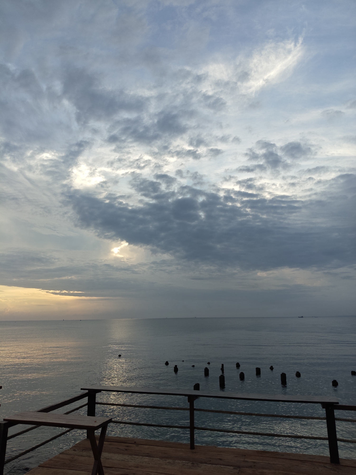 The image depicts a serene coastal scene at sunrise or sunset, likely in Lhokseumawe, Indonesia. In the foreground, a wooden deck with a railing provides a vantage point over the calm sea. A small, foldable wooden table is set up on the deck. The sea extends to the horizon, with several dark, weathered wooden pilings or remnants of a pier protruding from the water. The sky is filled with a mix of dramatic clouds, with patches of sunlight breaking through, casting a warm glow on the water's surface. A few distant boats are visible on the horizon, suggesting maritime activity. The overall atmosphere is tranquil and atmospheric, highlighting the natural beauty of the coastal landscape.