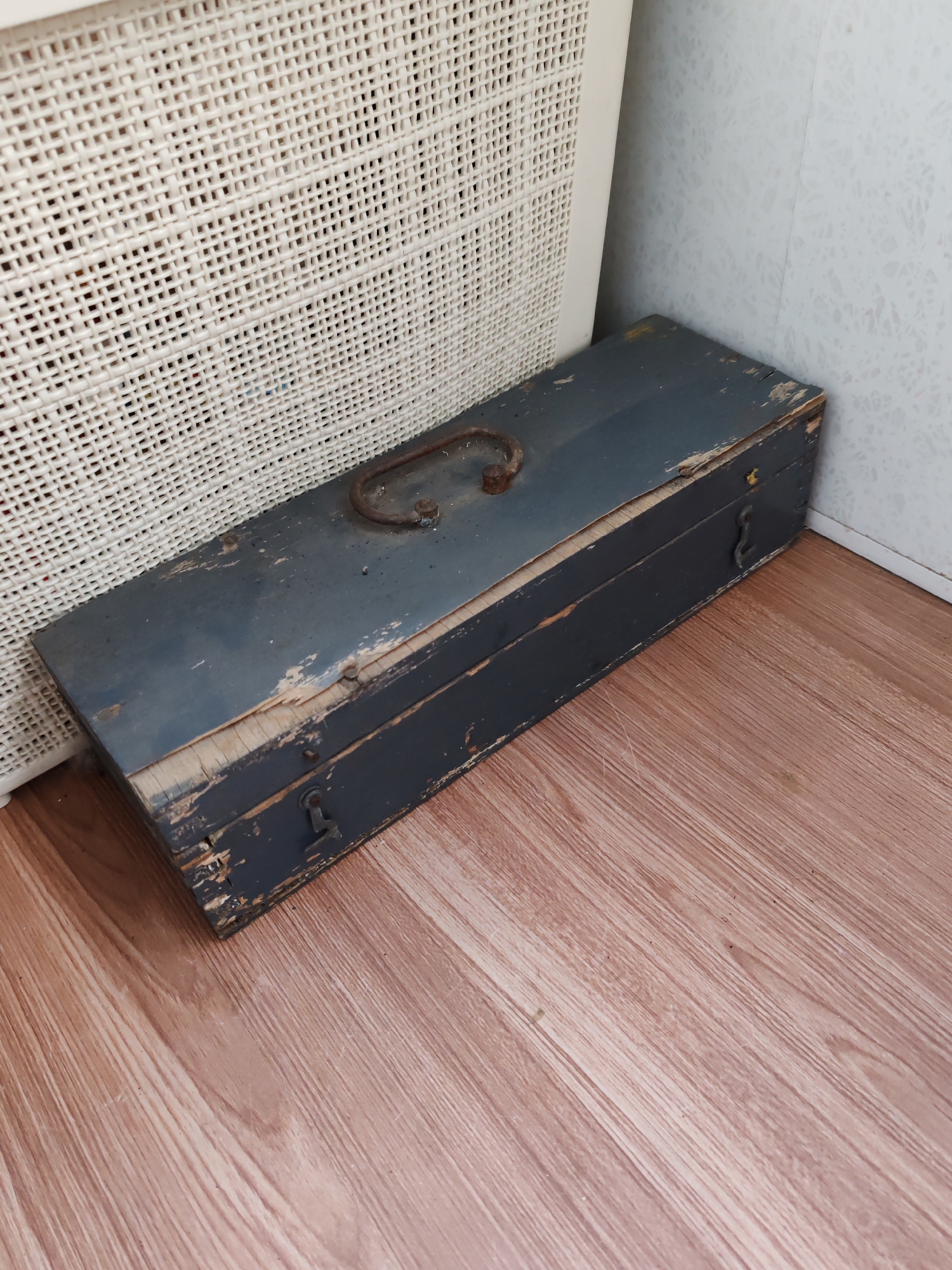 The image shows a weathered, blue-gray toolbox resting on a wood-patterned floor. The toolbox is rectangular and appears to be made of wood with a metal handle on top. It has visible signs of wear and tear, including chipped paint and exposed wood. The toolbox is positioned near a white, woven material object and a plain, light-colored wall. The environment suggests an indoor setting. The location is not discernible from the image.