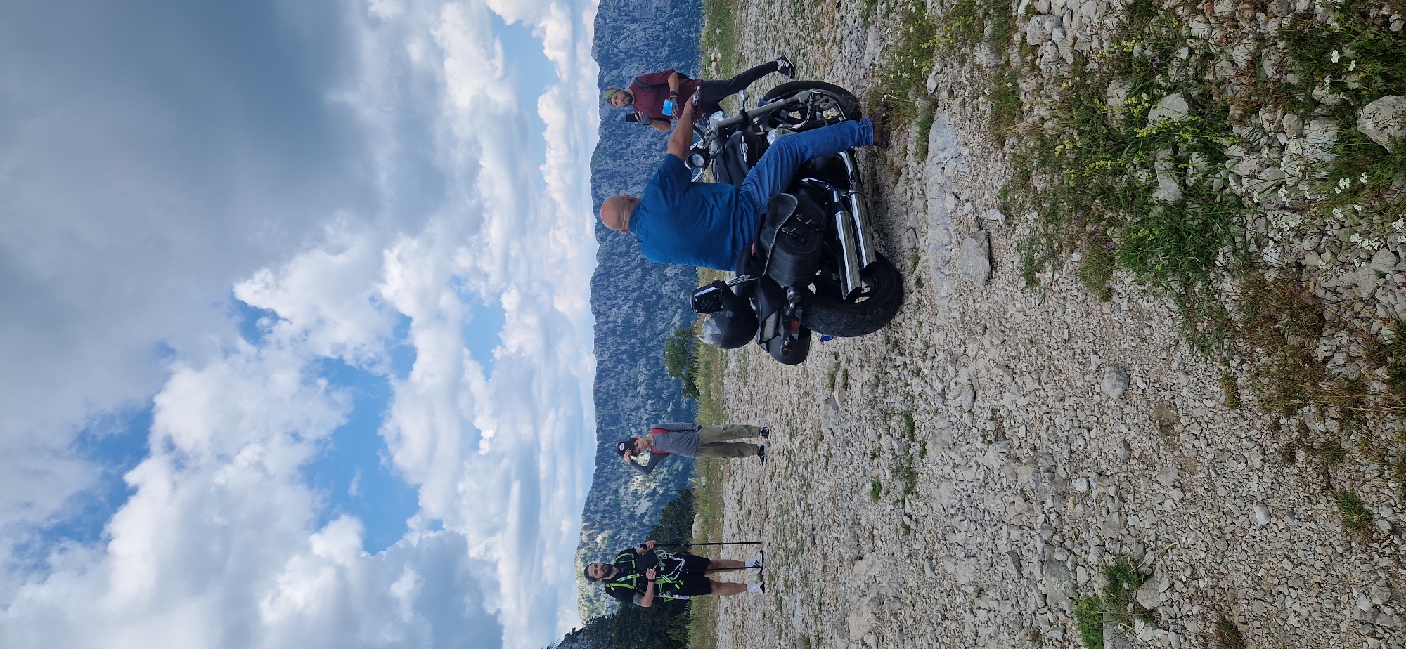 The image depicts a mountainous outdoor scene, likely near Dragalj, Montenegro, under a cloudy sky. In the foreground, a person is seated on a dark-colored motorcycle. To the left, another person wearing a harness and holding trekking poles stands on a rocky terrain. Between them, a young person in a grey shirt and cap is looking away from the camera. Further to the right, a person in a maroon shirt and a beanie holds up a mobile phone. The background features a rugged mountain range covered in trees, with a vast expanse of sky above dotted with clouds. The ground is covered with stones and some sparse vegetation.