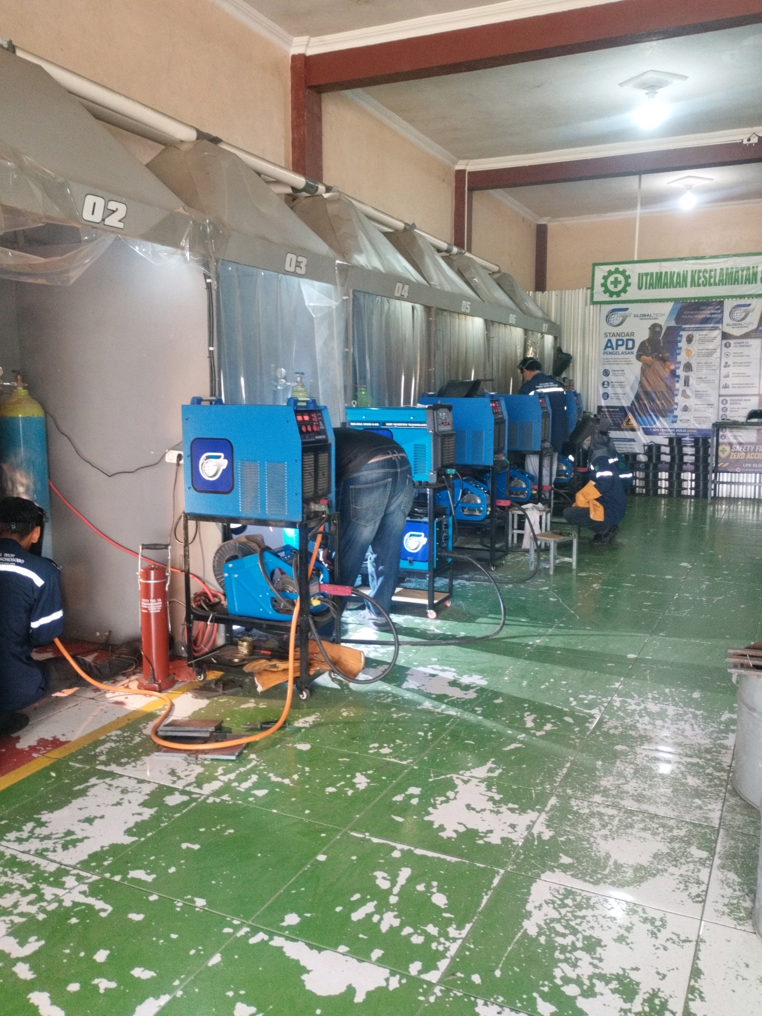 The image depicts a welding workshop, likely located in Selomerto, Indonesia, given the context. The environment features a series of welding stations, each equipped with a bright blue welding machine, protective curtains, and ventilation hoods overhead, marked with numbers 02, 03, 04, and 05. The floor is tiled in green with significant peeling white paint, indicating wear and tear.
Several individuals are present, all engaged in or preparing for welding activities. One person in a dark blue uniform is working near a welding machine on the left. Another individual, also in a dark blue uniform, is seen from the waist down, wearing jeans and working with a welding machine in the center. A third person, in a dark blue uniform with reflective stripes, is kneeling and attending to a welding machine on the right. A fourth person in a similar uniform is visible further back.
Prominently displayed on the wall is a green banner with white text that reads "UTAMAKAN KESELAMATAN KERJA" (Prioritize Work Safety) and a poster detailing "STANDAR APD PENGELASAN" (Welding PPE Standards), featuring illustrations of safety equipment. Another sign in the background advertises "SAFETY FIRST ZERO ACCIDENT".
Other objects in the scene include gas cylinders (one blue and one yellow), welding cables, welding tools, and a small stool. The overall impression is a functional, albeit somewhat cluttered, industrial training or operational space focused on welding.