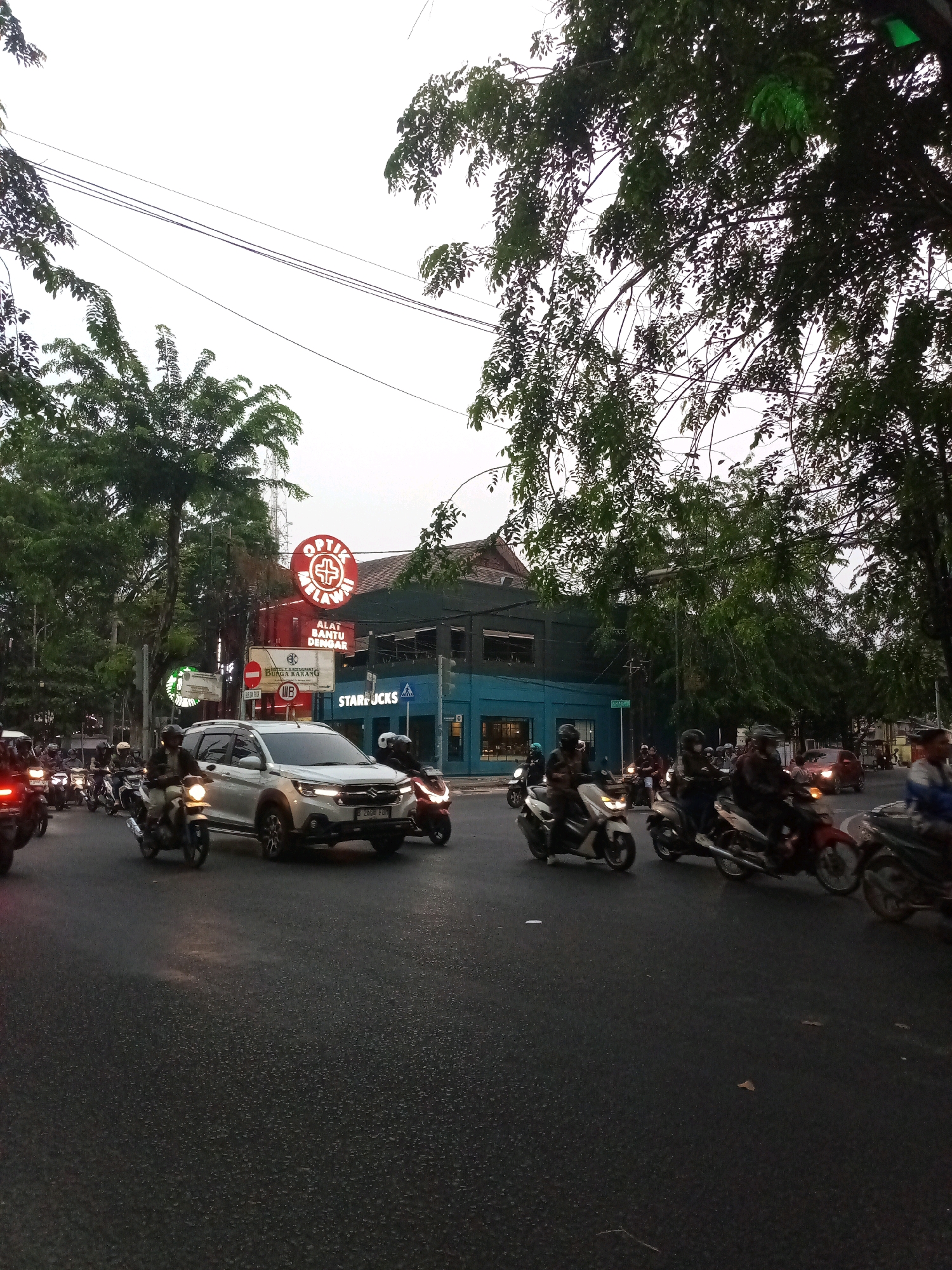 The image shows a street scene in the city of Bekasi, Indonesia. Several motorcycles and a white SUV are on the road, suggesting moderate traffic. The vehicles are moving or stopped at what appears to be an intersection. Buildings are visible along the street, including a prominent building with a red circular sign that reads "OPTIC MELAWAY" in white lettering. Below this sign, there is text that says "ALAT BANTU DENAR". Another building has a Starbucks logo. Trees with dense green foliage line the street, and power lines are visible overhead. The sky is overcast, indicating potentially low light conditions, possibly dawn or dusk. The road surface is asphalt and appears wet in some areas, suggesting recent rain.