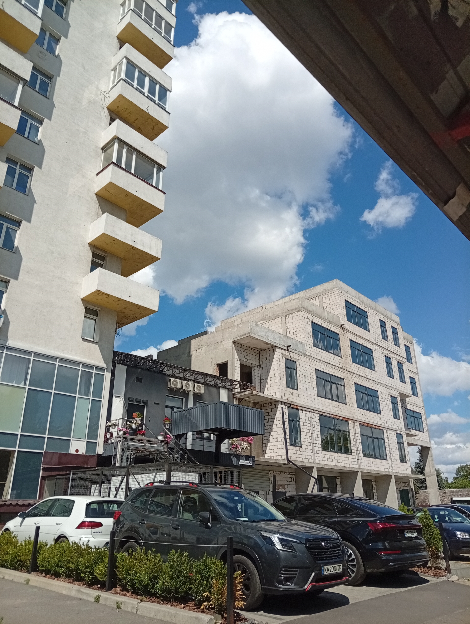 This is a photograph taken outdoors on a sunny day with scattered clouds. The scene depicts a street with several modern buildings and parked cars.
In the foreground, a row of neatly trimmed green bushes lines the edge of a sidewalk or parking area. Behind the bushes, several cars are parked. A dark gray SUV is prominently featured, with its front facing left. To its left, a white hatchback is visible. Further to the right, a black sedan is parked, followed by another dark-colored car, possibly a sedan or hatchback.
Behind the cars, there are several buildings. On the left, a tall, light-colored building with many windows and protruding balconies is visible. The balconies have yellow undersides and appear to be enclosed with glass. To the right of this building, there is a lower, dark gray building with prominent air conditioning units mounted on its exterior. Above this dark gray building, a multi-story building constructed with white bricks is under construction or recently completed, featuring large, dark-framed windows. The sky above is a bright blue with fluffy white clouds.
The overall impression is a contemporary urban or suburban setting. The sunlight suggests it is daytime, likely mid-morning or afternoon. There are no people visible in the photograph. The scene appears calm and ordinary, with no specific event taking place.