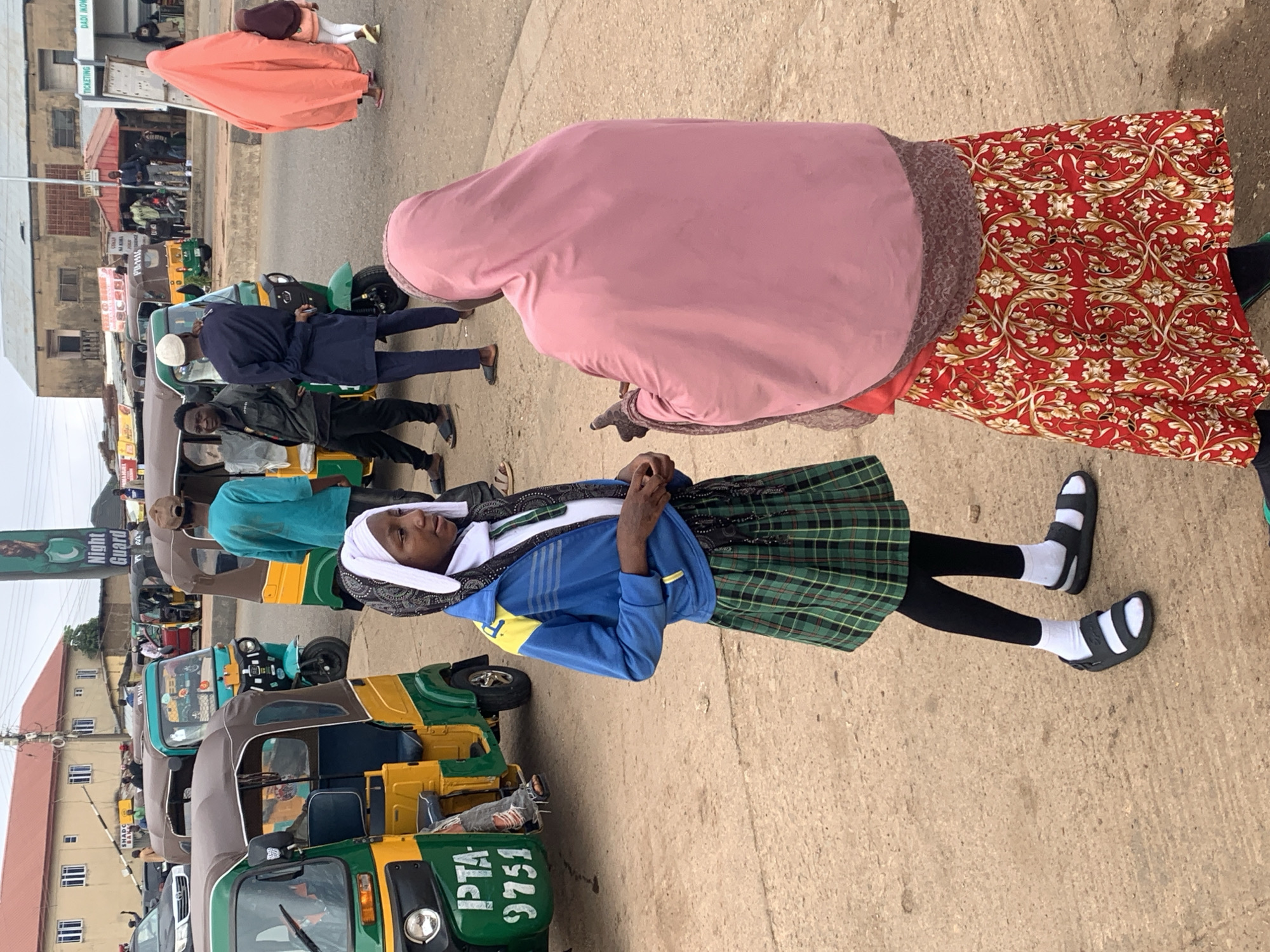 A medium shot captures a street scene in Gura Top, Nigeria, likely a transportation hub. In the foreground, two women face each other, engaged in conversation. The woman on the left, a young girl wearing a white headscarf, blue and yellow jacket, white shirt, black leggings, white socks, and green plaid skirt, stands with her hands clasped. The woman on the right, veiled in a pink hijab and a patterned red skirt, gestures with her left hand. Behind them, several auto-rickshaws, painted in vibrant green and yellow, are parked. One rickshaw in the left foreground displays the license plate "PTA 9751." In the background, a banner with the text "Night Guard" and an image of a man is visible. Buildings with red roofs line the street, and other people are seen in the distance, some near the rickshaws. The ground is a mixture of sand and concrete.